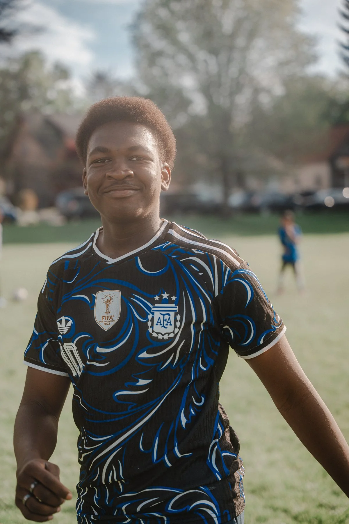 Young man wearing a black and blue soccer jersey with FIFA World Cup 2022 and Argentina Football Association logos, smiling outdoors on a field.