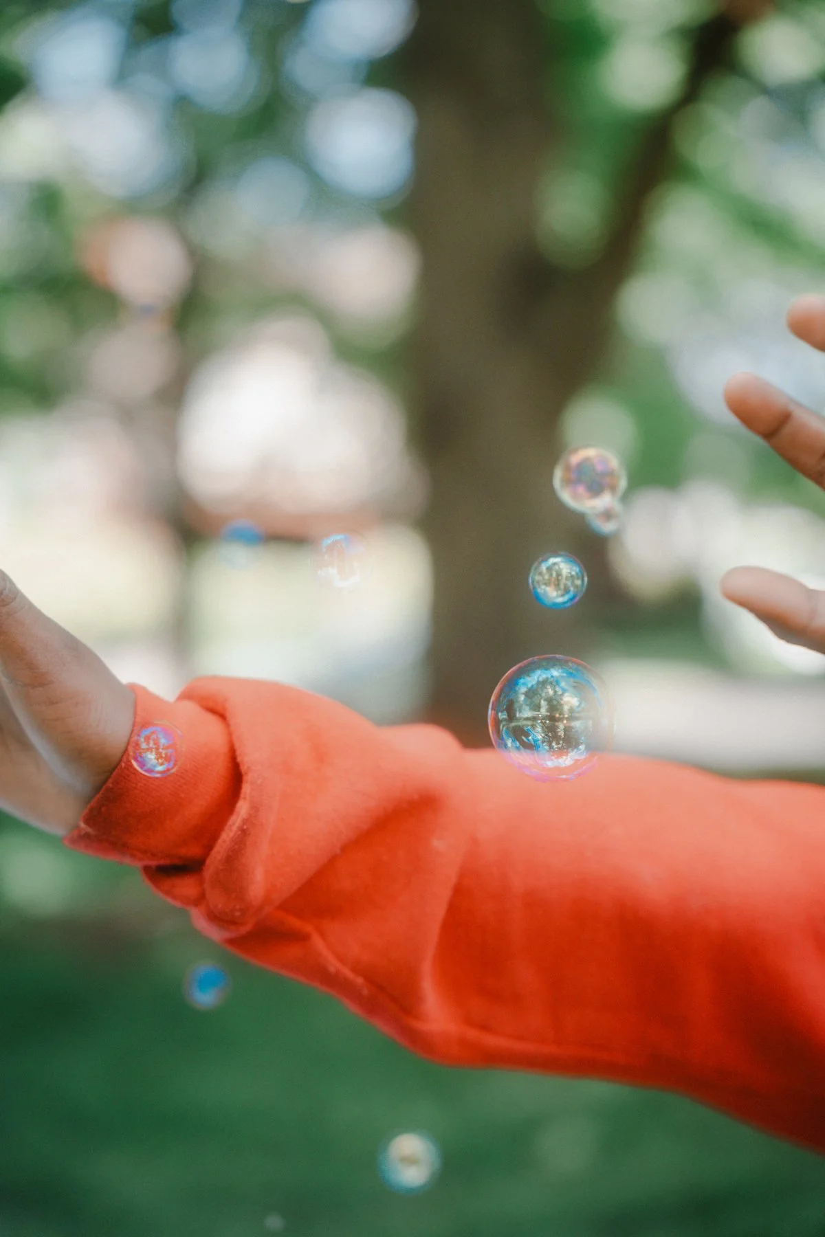 Close-up of a person's hand with a red sleeve reaching out towards floating soap bubbles outdoors, with a blurred tree in the background.