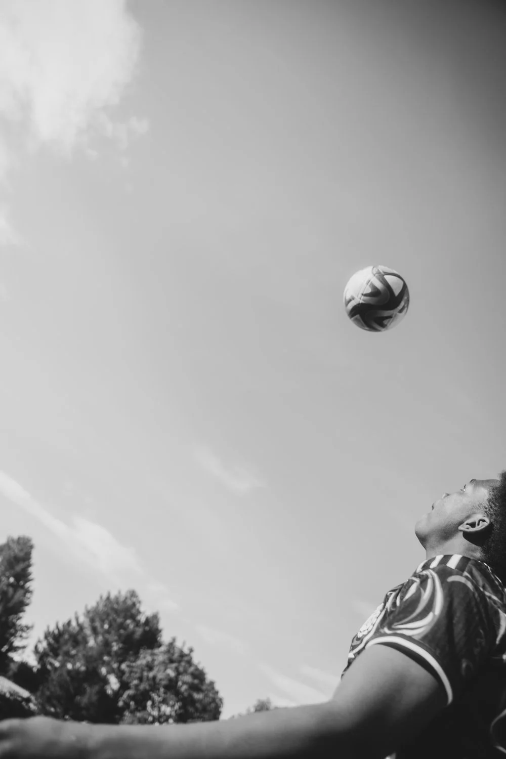 A young man leans back and looks up at a soccer ball in the sky outdoors, with trees and clouds in the background.