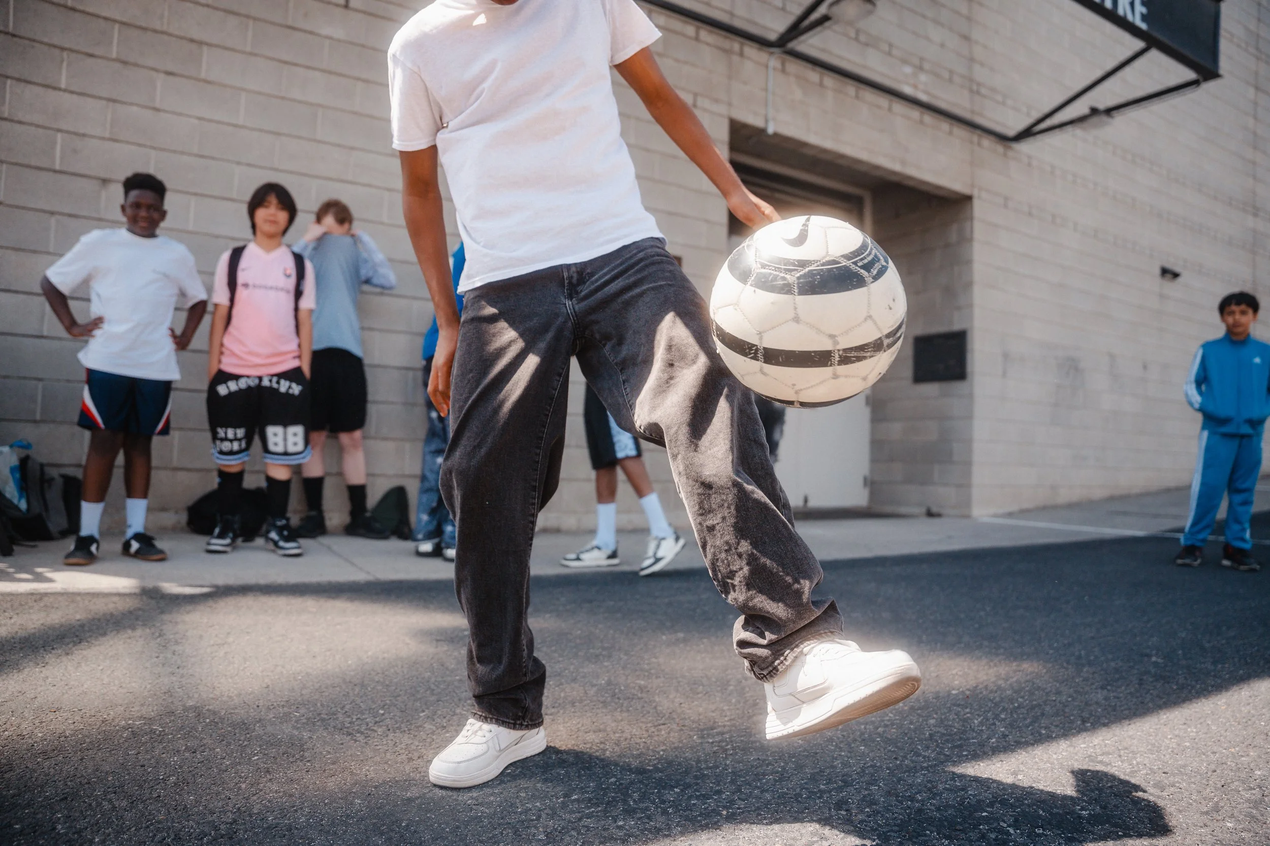 A person in a white t-shirt and dark jeans is playing with a soccer ball on the street, while a group of children watches nearby against a brick wall.