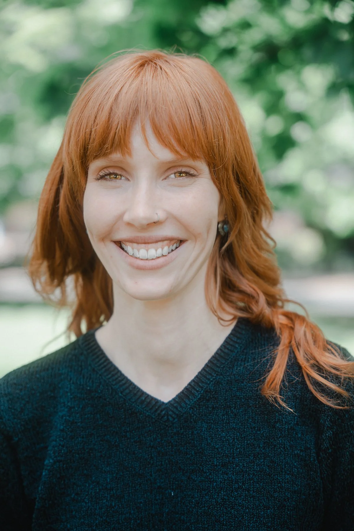 A smiling woman with red hair, wearing a black sweater, standing outdoors with a blurred green background.