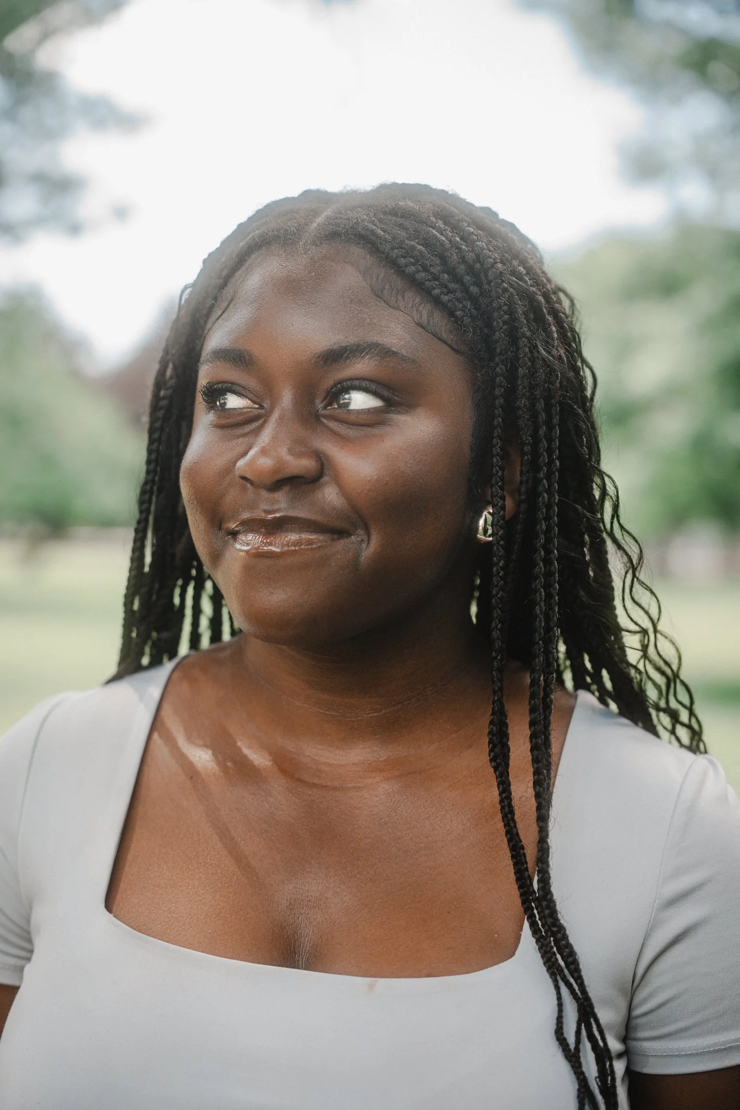 Close-up of an African American woman with long braided hair, wearing a light gray top with a wide neckline, outdoors with blurred green trees and sky in the background.