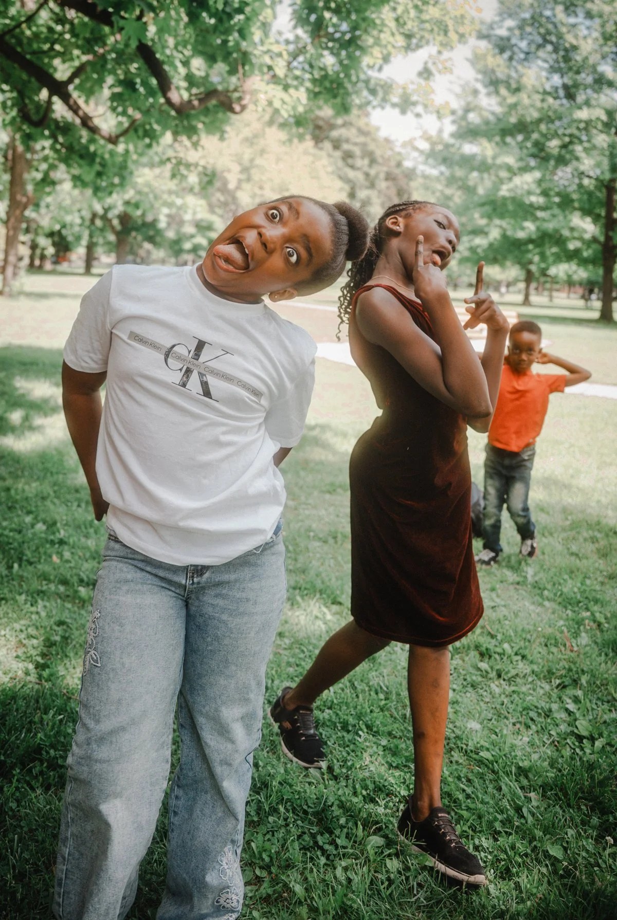 Three children playing outdoors in a park with green grass and trees. One girl in the foreground is making a silly face with her tongue out, wearing a white Calvin Klein T-shirt and jeans. A girl behind her is striking a playful pose with her hand near her face, wearing a dark dress. A young boy in the background is listening with earphones, wearing an orange shirt and jeans.