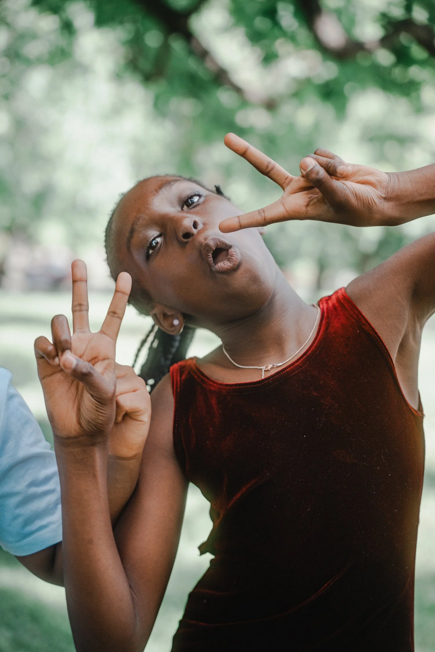 A young girl with braids and earrings making peace signs with her fingers while looking at the camera. She is wearing a red velvety top and a necklace, with a background of green trees.