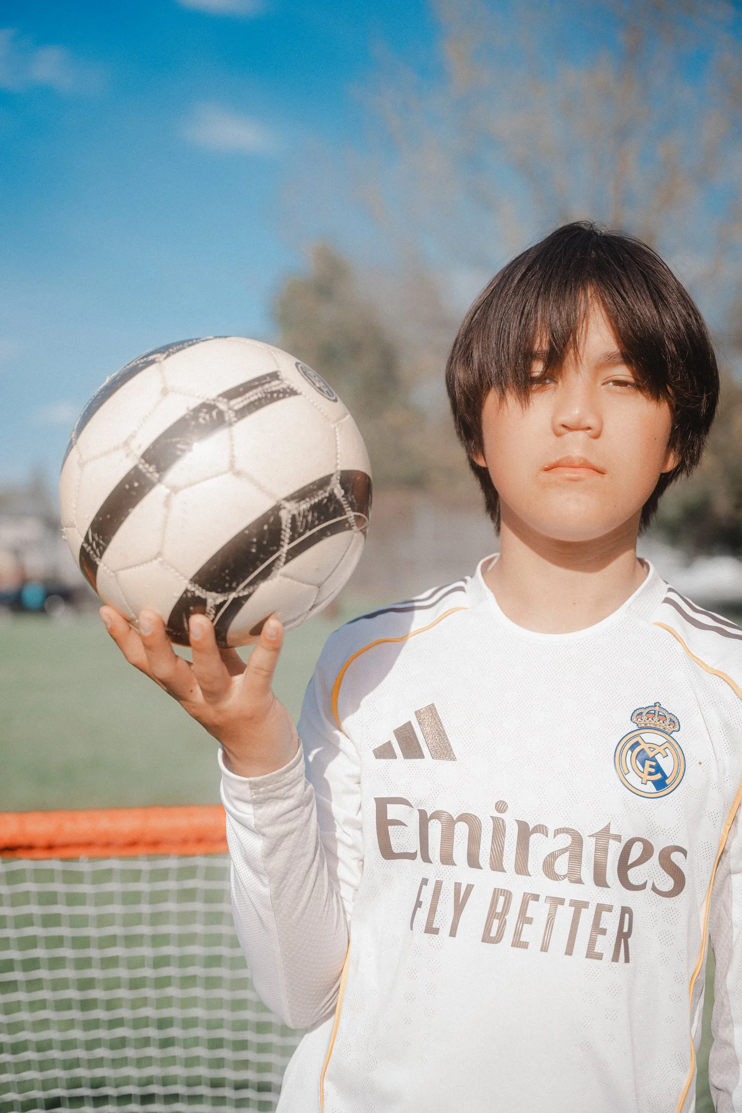 A young boy with dark hair wearing a white soccer jersey with the Real Madrid logo and Adidas branding, holding a black and white soccer ball, standing on a soccer field with a goal and trees in the background on a sunny day.