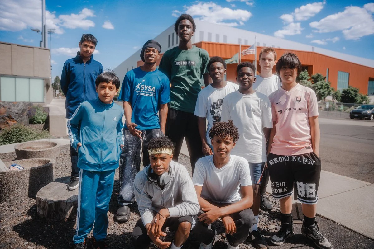 Group of young boys and teens standing outside on a sunny day in front of a modern building, some wearing sports clothing and headphones, with a clear blue sky overhead.
