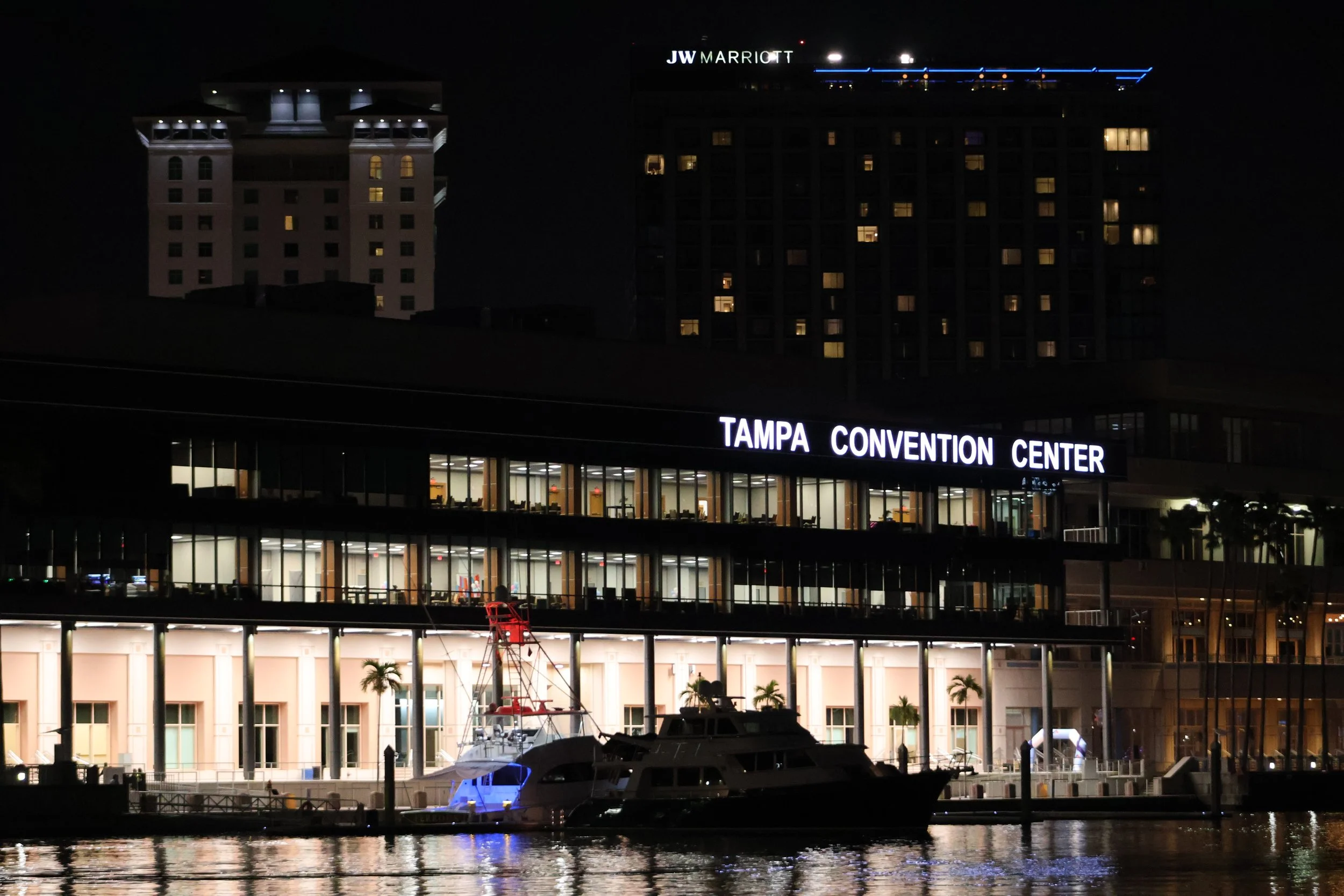 Night view of the Tampa Convention Center with illuminated buildings, boats docked in the water, and the JW Marriott hotel in the background.