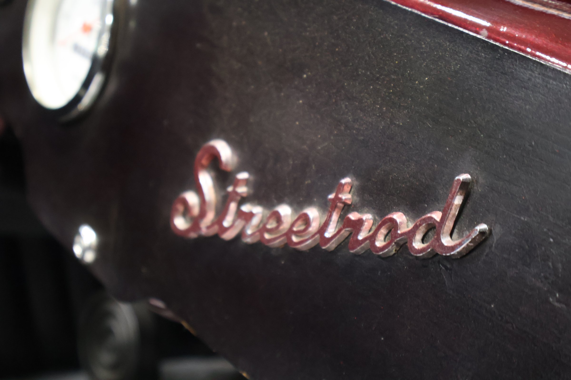 Close-up of a metallic badge with the word "Standard" in cursive, mounted on a black surface with a faint red edge visible in the background.