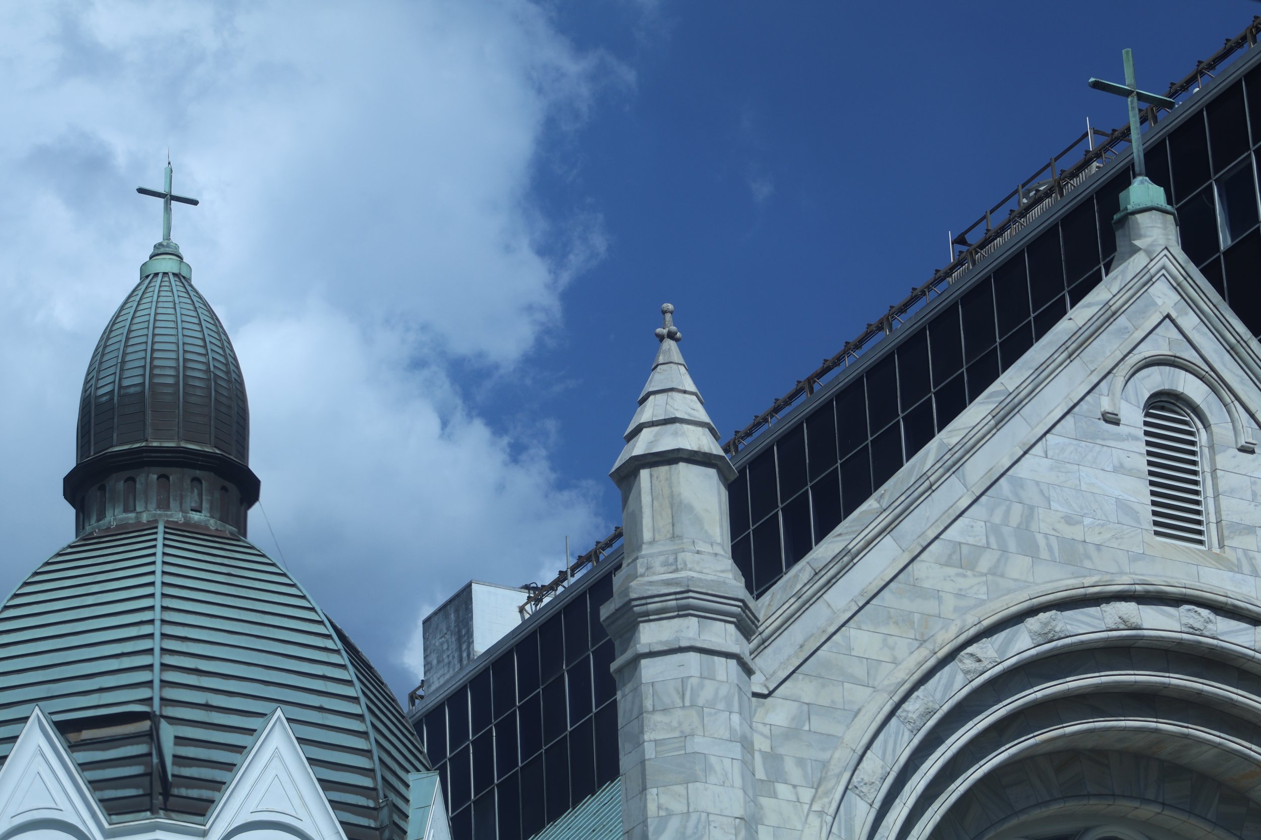 Close-up of a church's roof with gray and white stonework, stained glass windows, and a cross at the top of the steeple against a partly cloudy sky.