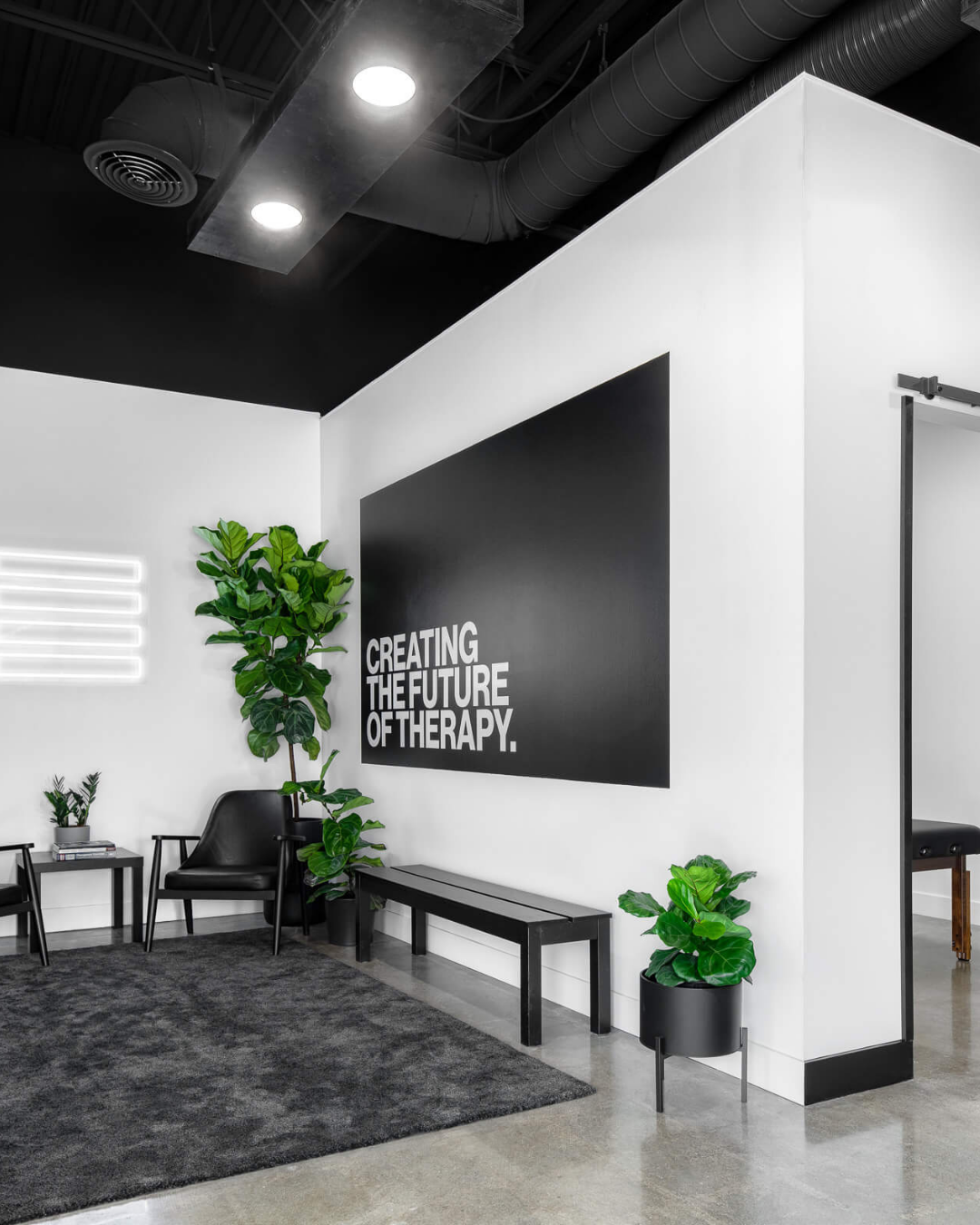 Modern office reception area with black and white decor, geometric black wall sign reading "Creating the future of therapy," black chairs, benches, potted plants, and neon wall art.