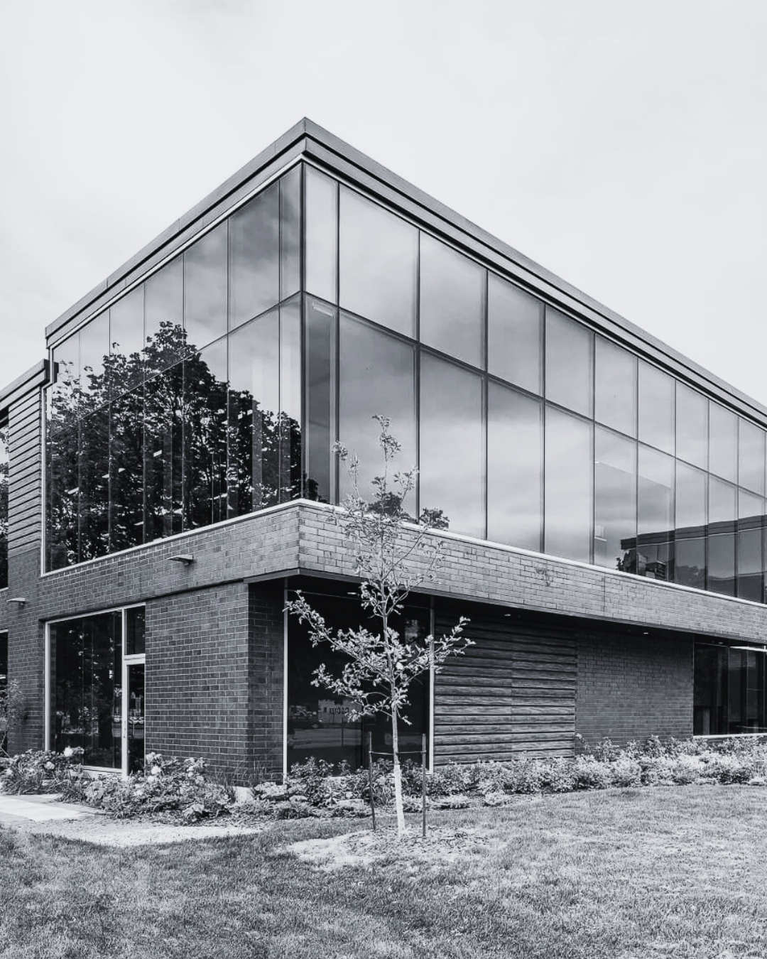 Modern two-story building with large glass windows, brick and siding exterior, surrounded by grass and small trees, reflected clouds in the windows.