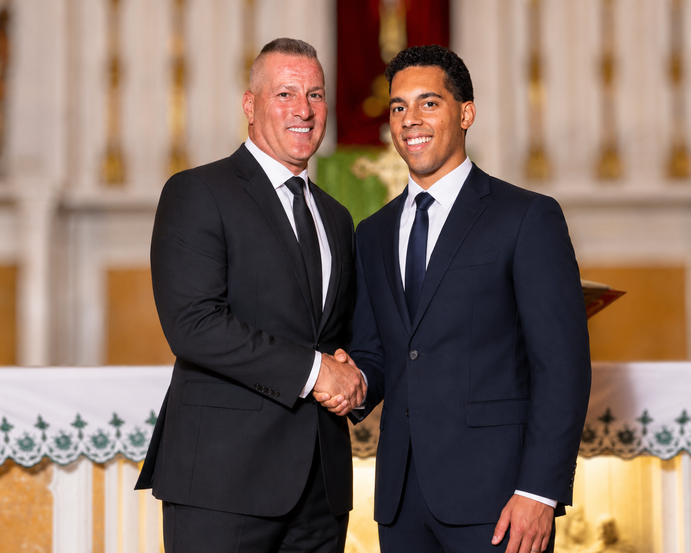 Two men in suits shaking hands inside a church or chapel, with an altar and religious decorations in the background.