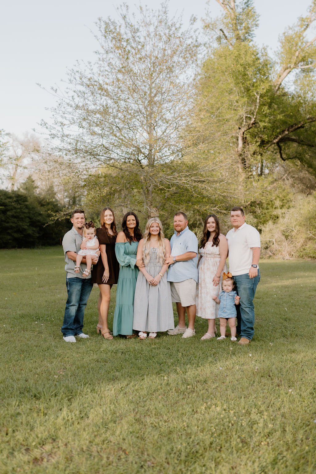 A family of eight people standing on a grassy field with trees in the background, smiling at the camera during an outdoor photo shoot.