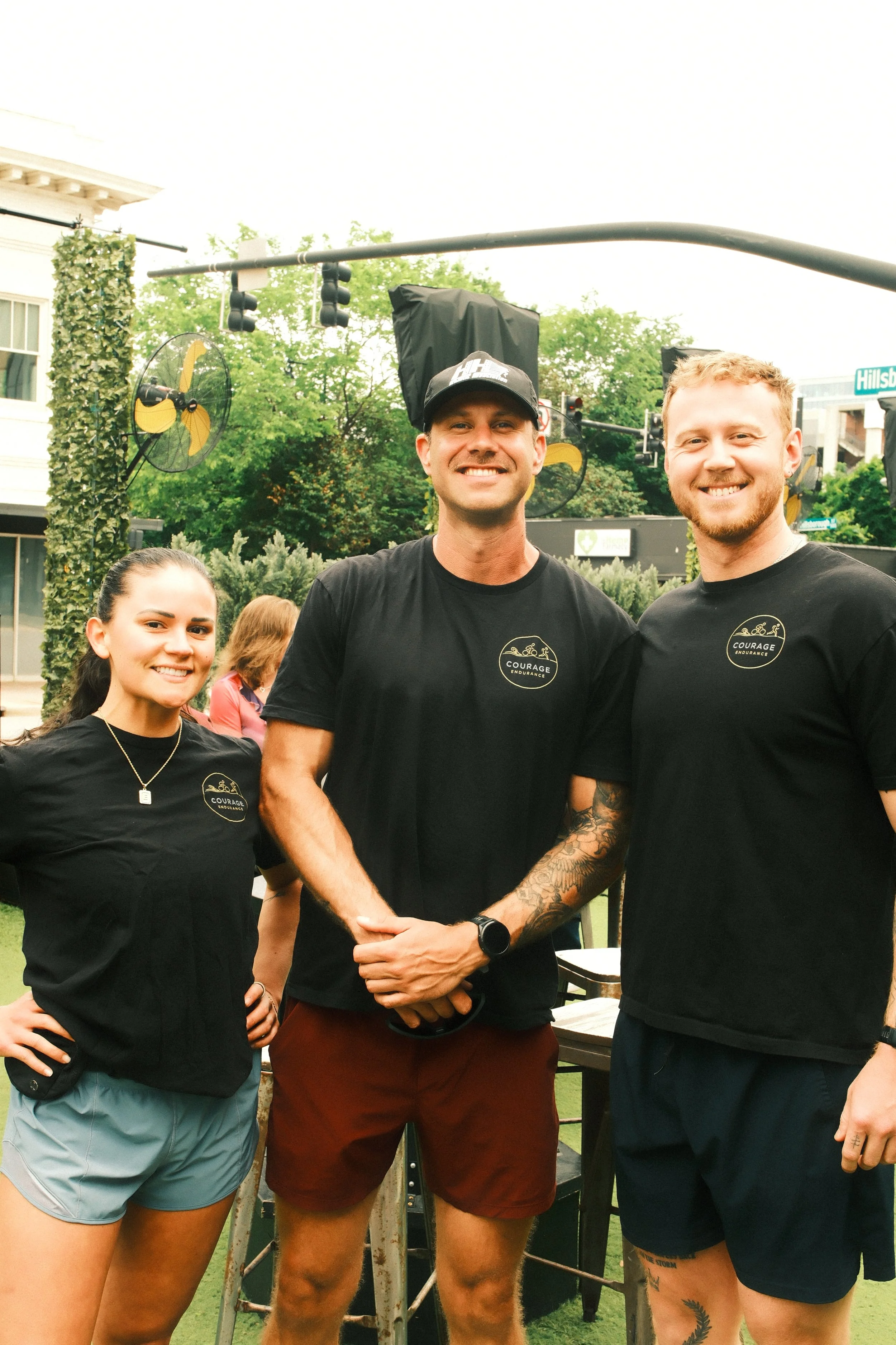 Three smiling people standing outdoors in front of a stage with trees and buildings in the background. They are wearing black t-shirts, and one person has tattoos.