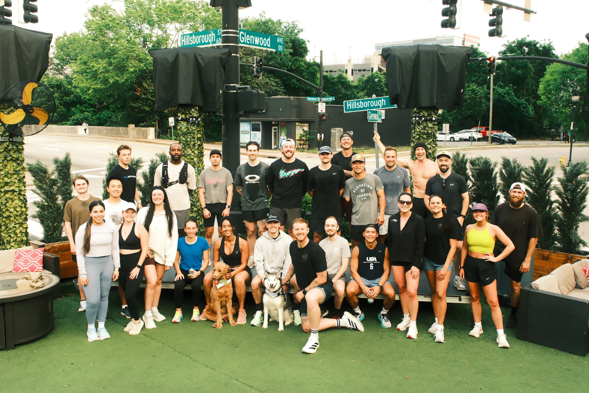 Group of smiling people, some with dogs, gathered outdoors near street signs for Hillsborough Street and Glenwood Avenue in an urban area with trees, cars, and buildings in the background.
