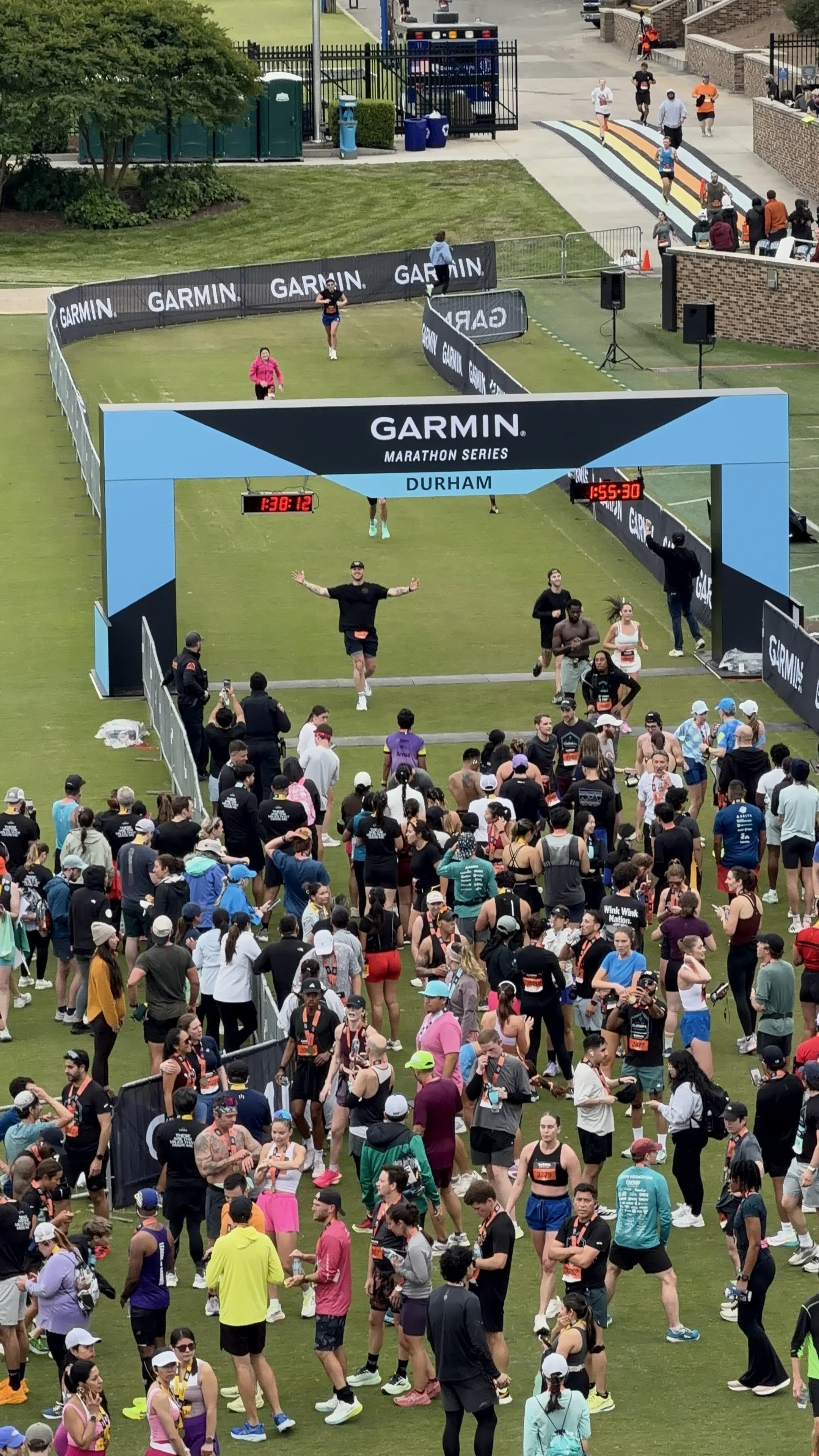 People gather at finish line of a marathon in Durham, with a large crowd of runners and spectators, some crossing the line and others waiting nearby. The finish line has a blue and black Garmin Marathon Series banner and digital timers.