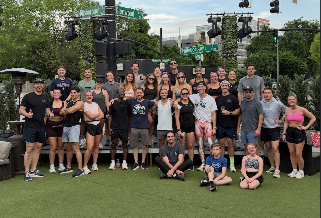A group of people gathered outdoors on a patio with greenery and city buildings in the background. They are standing and sitting, posing for a photo beneath street signs for Hillsborough Street and Glenwood Avenue.