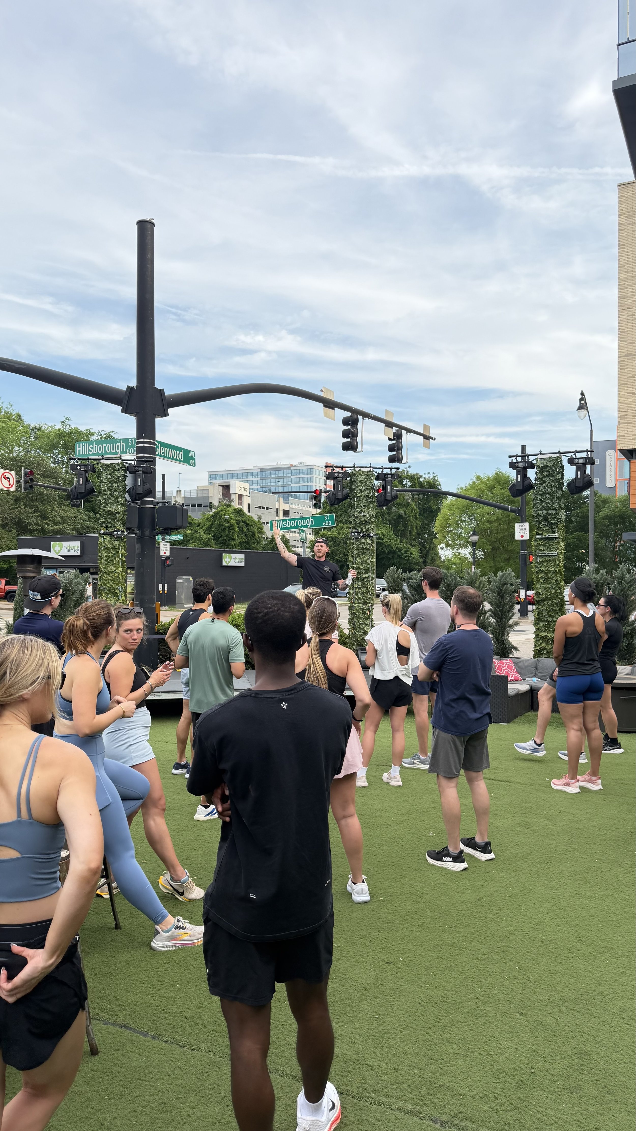 A group of people gathered outdoors in a park or plaza area, listening to a man speaking on a stage. The setting includes street signs for Hillsborough and Glenwood, traffic lights, and nearby buildings. The weather is partly cloudy.