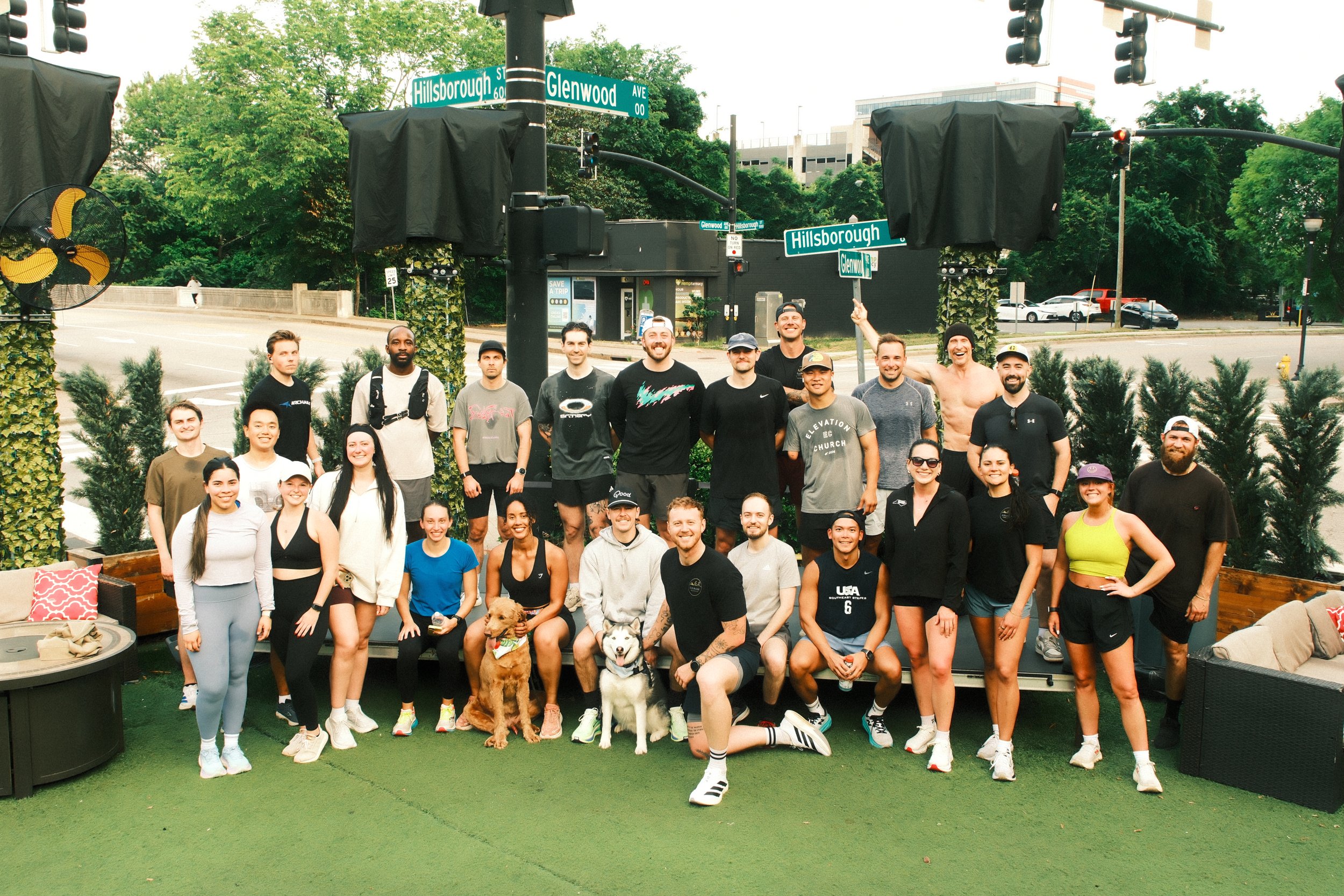 Large group of people and two dogs gathered outdoors near street signs for Hillsborough and Glenwood Ave, smiling and posing for a group photo.