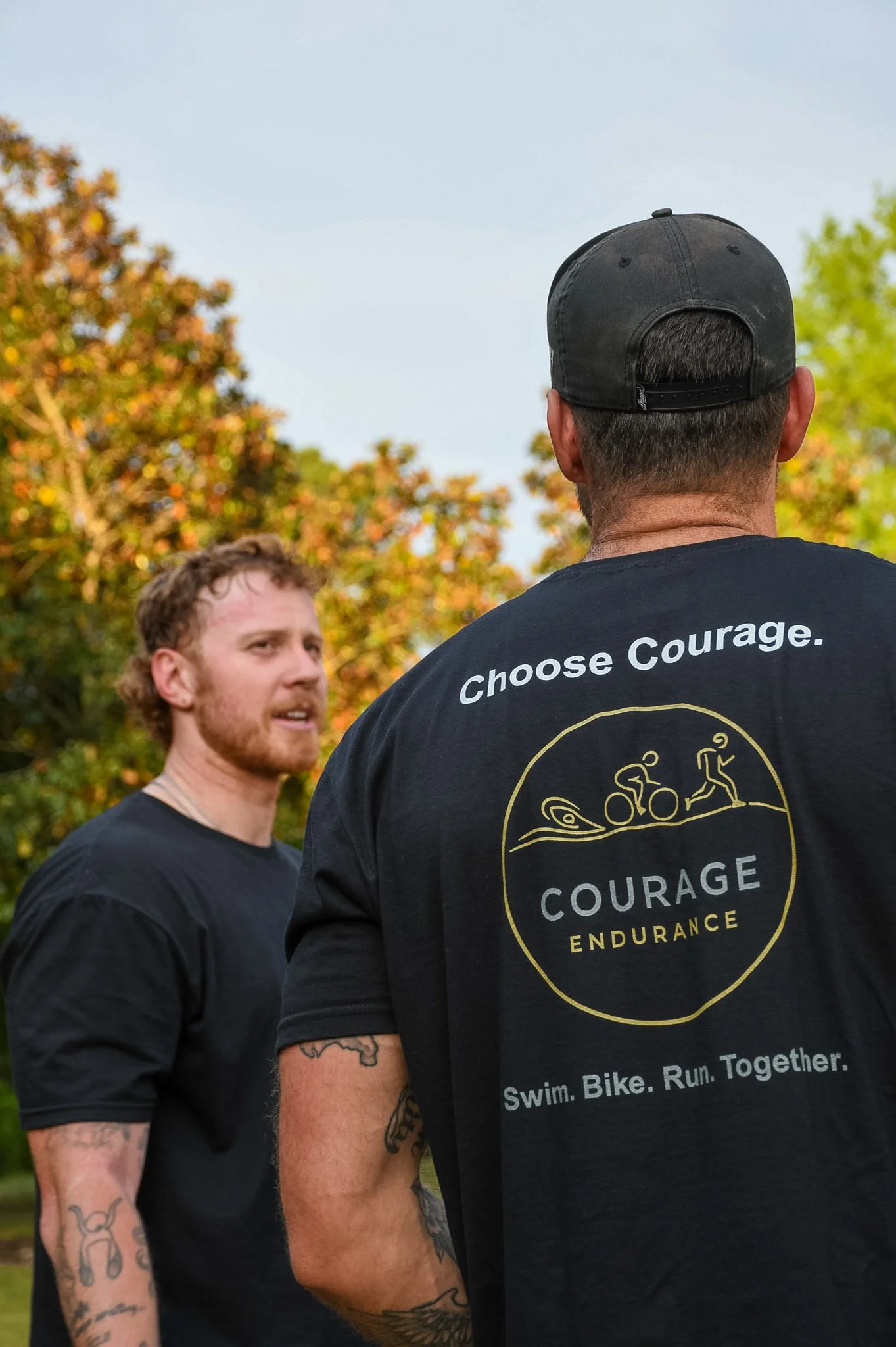 Two men standing outdoors in front of trees, one facing away wearing a black cap and shirt with motivational text, the other facing slightly to the left with a beard, tattoos on arms, and wearing a black T-shirt.