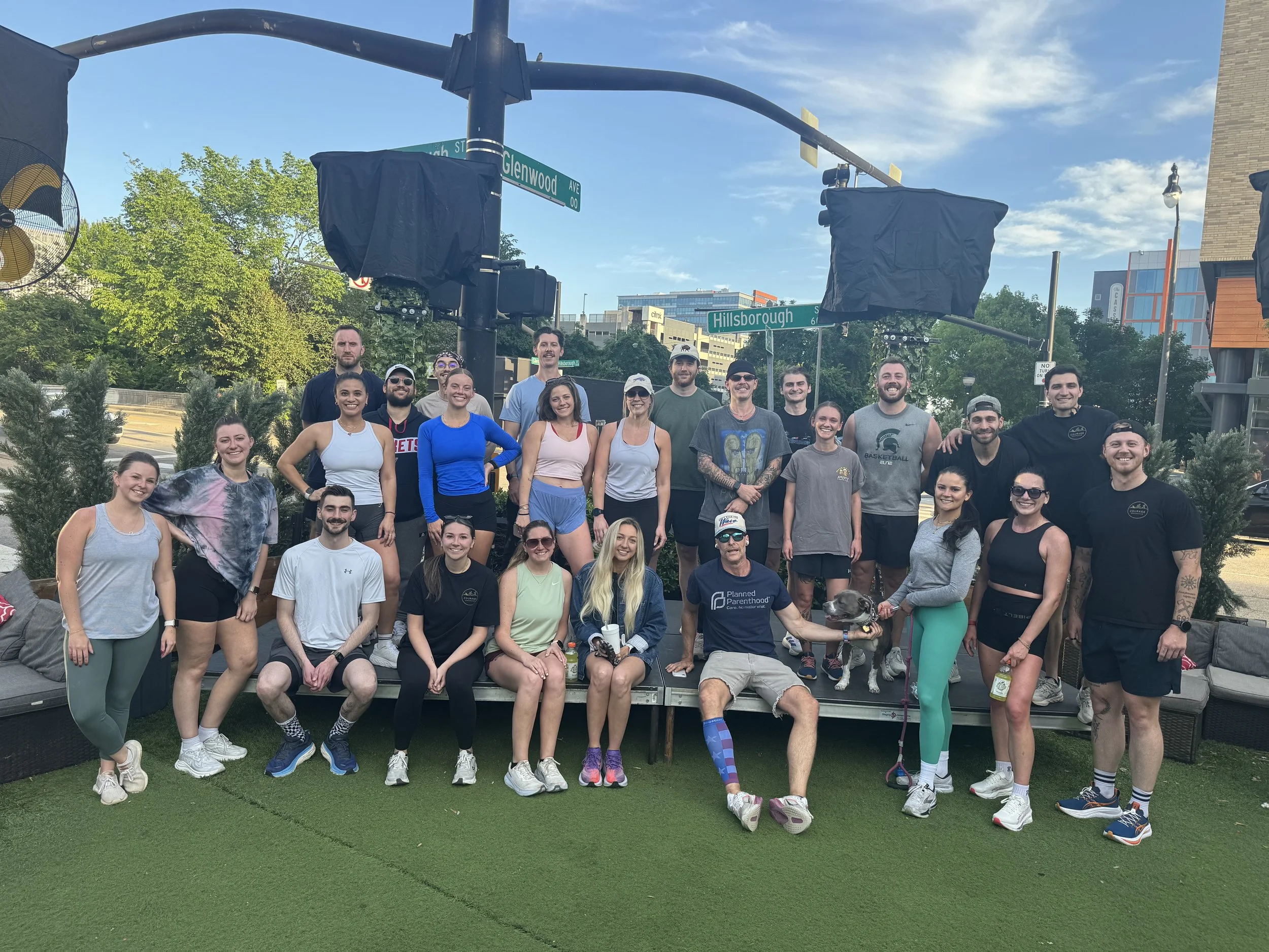 Group of people gathered outdoors on a green grass area, smiling for a photo, with some wearing athletic clothing.