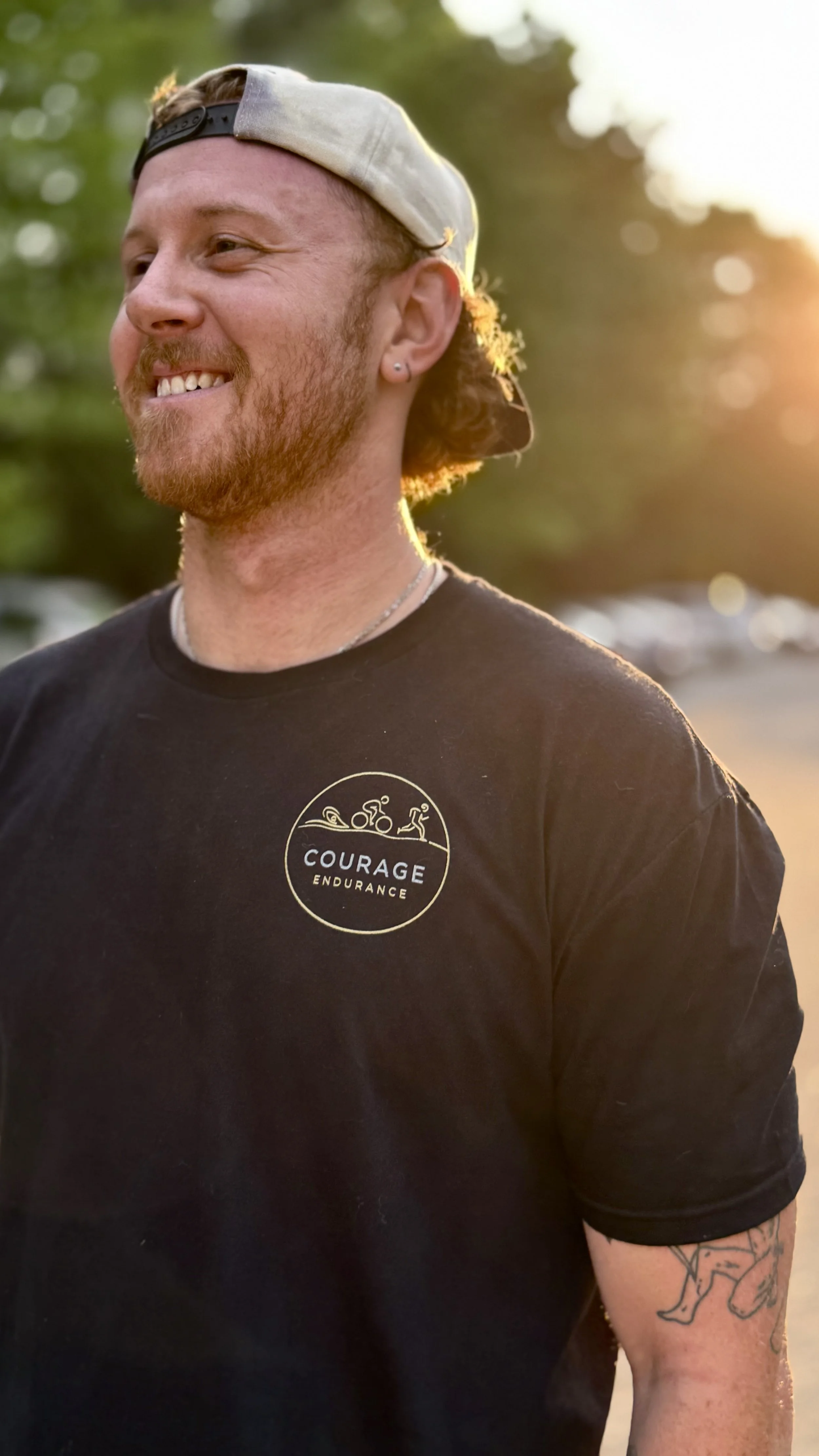 A man smiling outdoors at sunset, wearing a black T-shirt with a logo reading 'Courage Endurance' and a backward gray cap.