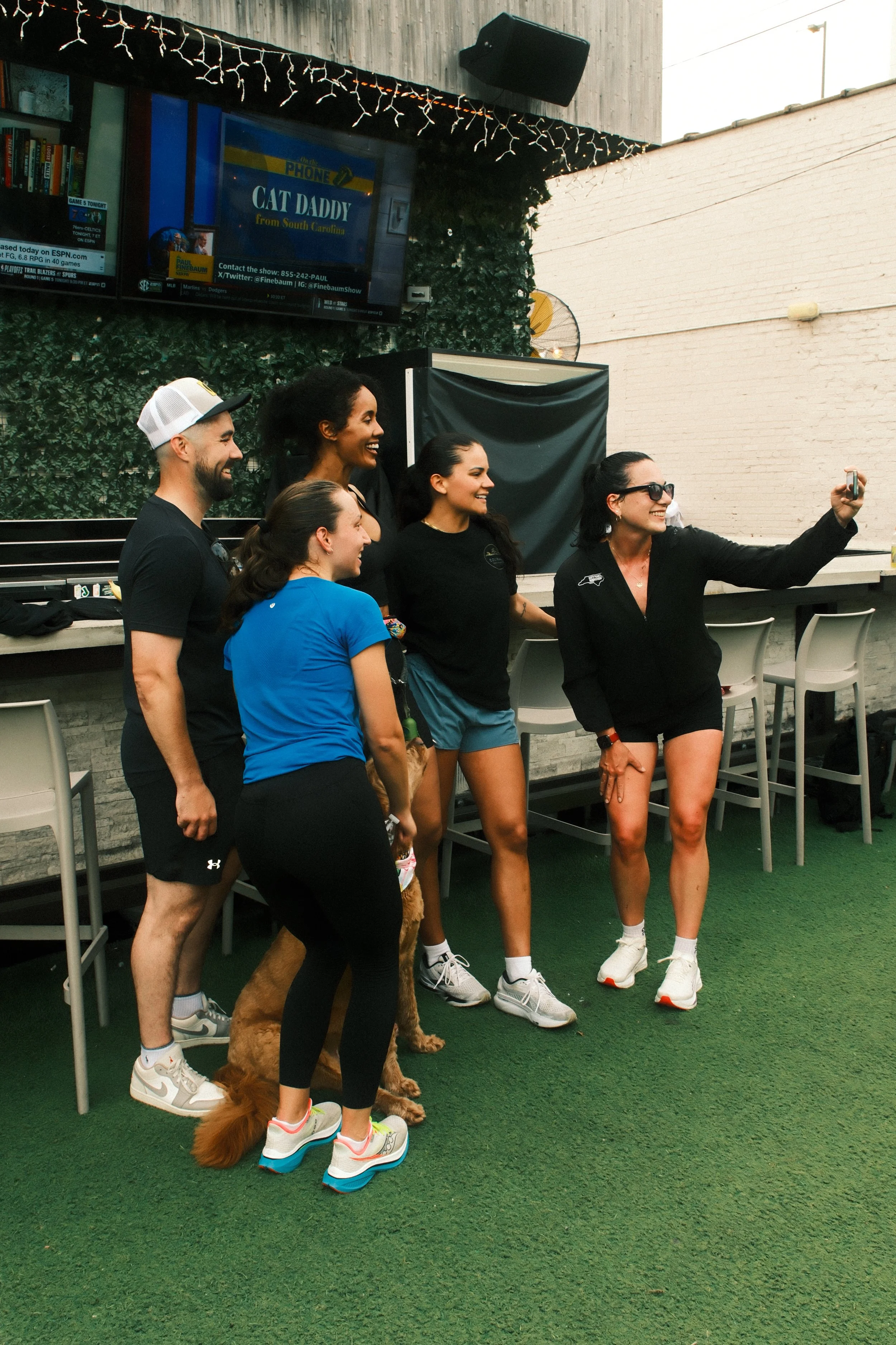 Group of five friends taking a selfie in an outdoor patio area, with a TV screen in the background displaying the show "Cat Daddy" from South Carolina, and string lights hanging along the top.