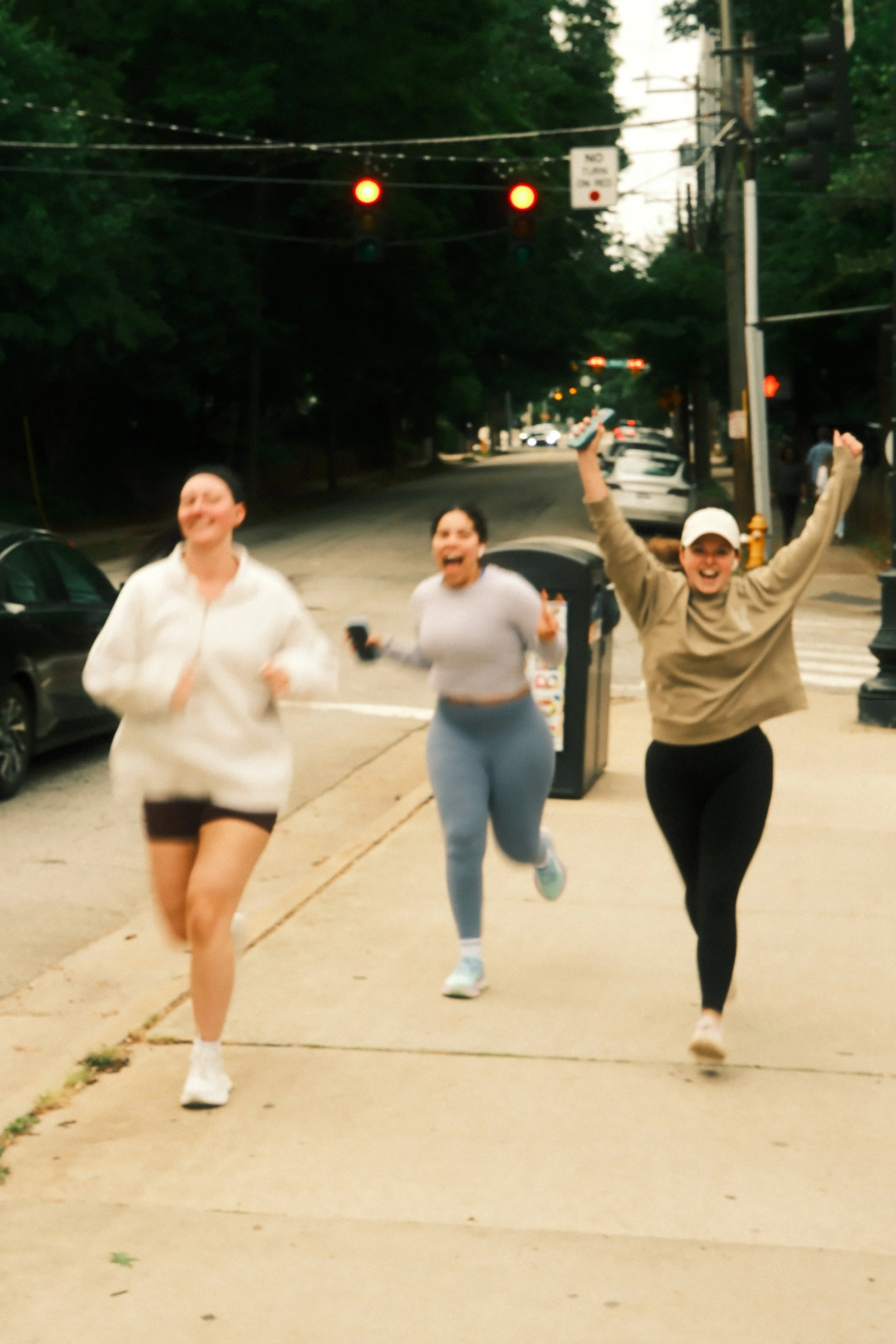 Three women running on a sidewalk with red traffic lights overhead and trees in the background.