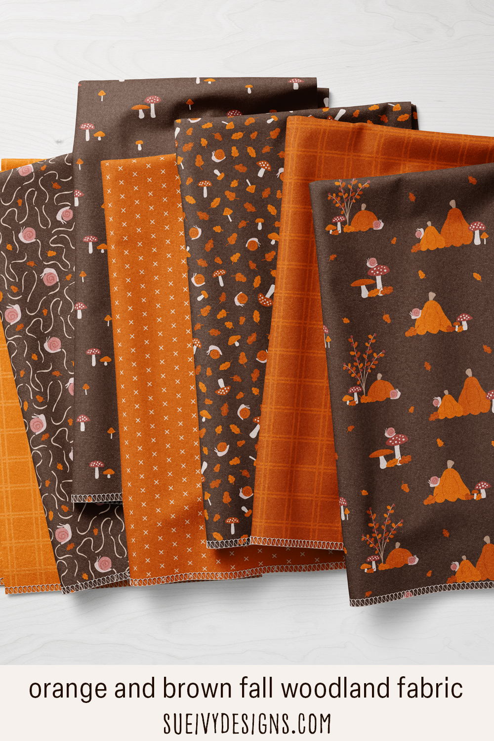 Snails & Mushrooms fabric fanned on pale wood — pumpkins, snails, mushrooms & leaves on coffee brown, plus plaid & cross blenders in orange.