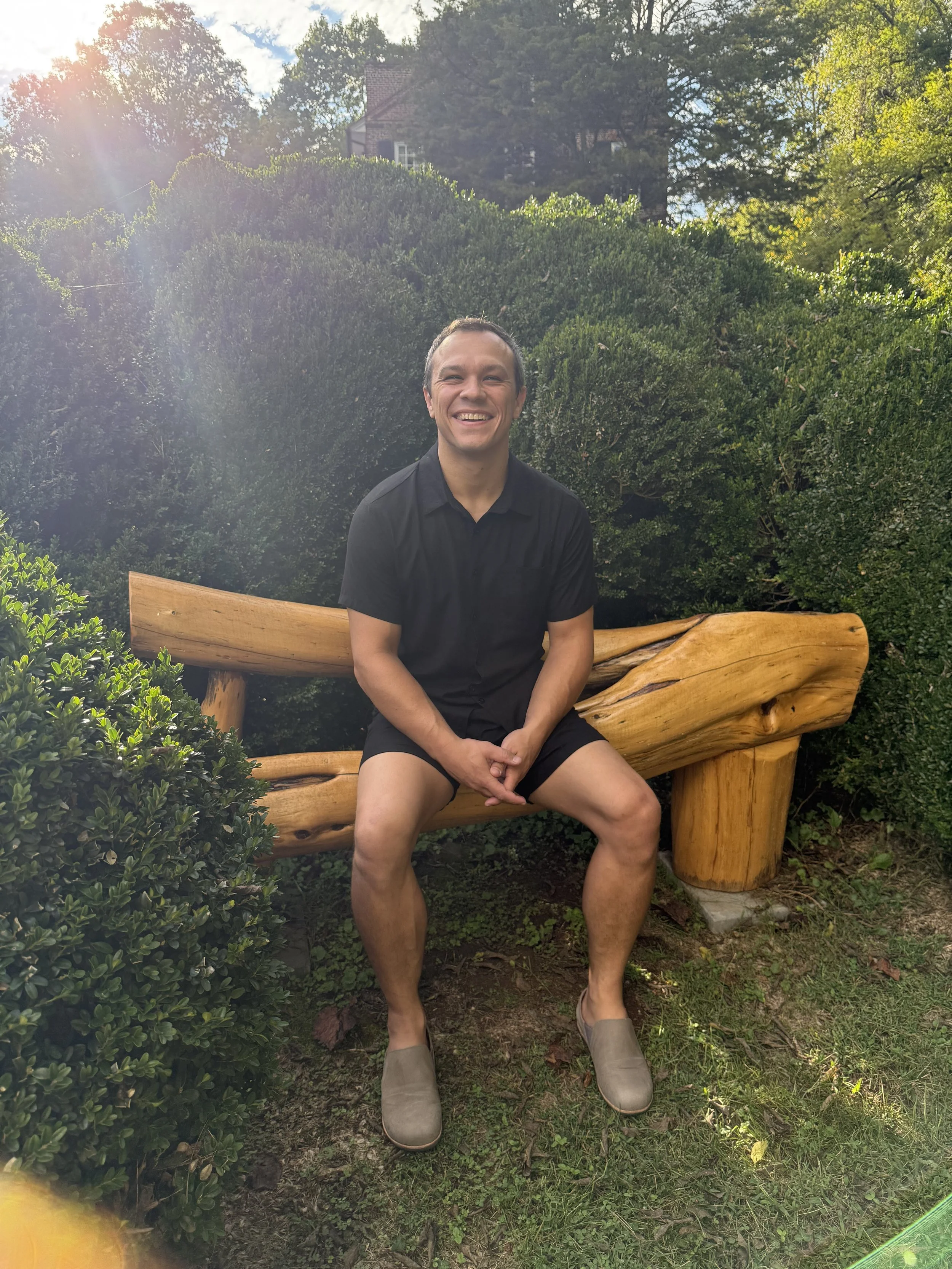 A smiling man sitting on a rustic wooden bench outdoors surrounded by greenery. Sunlight filters through trees in the background.