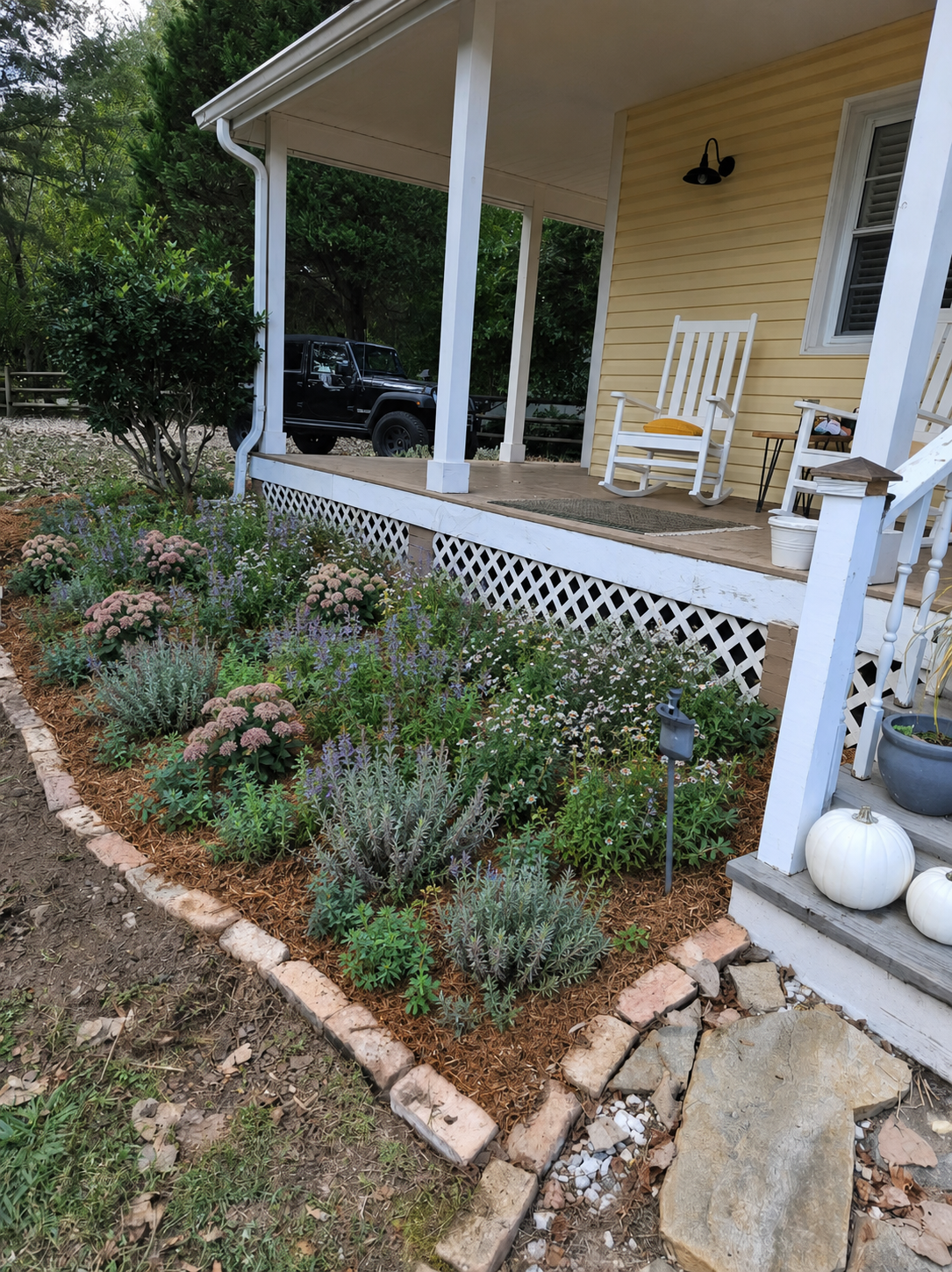 A front porch with yellow siding, white rocking chairs, and outdoor decor. A garden with various plants and flowers borders the porch. There are pumpkins and a potted plant near the stairs.