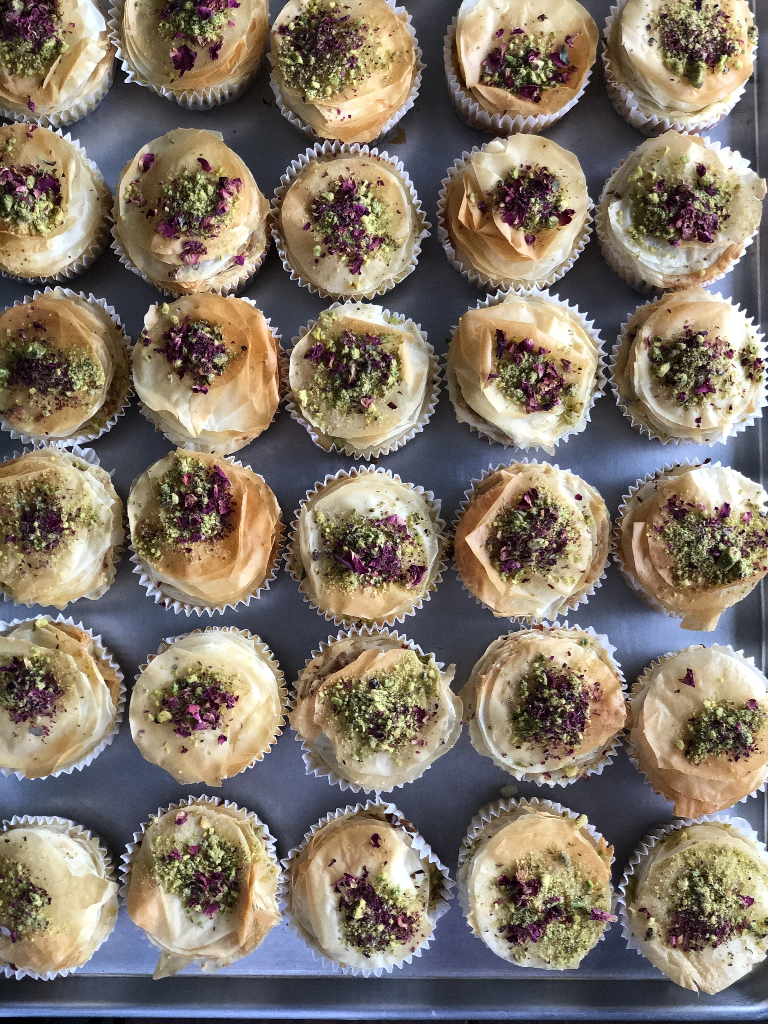 A tray of assorted cupcakes decorated with white and tan frosting, topped with purple and green edible flowers or crushed herbs.