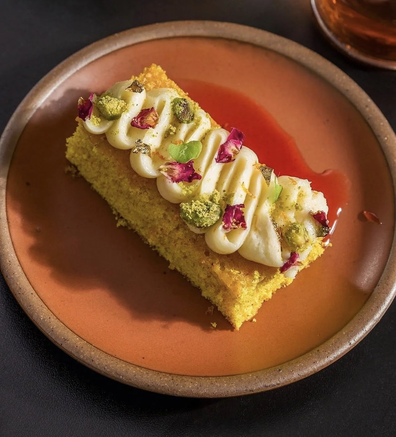 A rectangular slice of yellow cake topped with white icing and garnished with pink and green petals and green crushed pistachios on a brown ceramic plate.