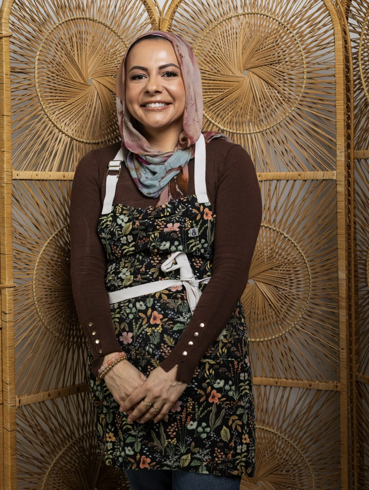 A smiling woman wearing a patterned apron and a pink and blue headscarf, standing in front of a decorative woven bamboo screen with circular patterns.