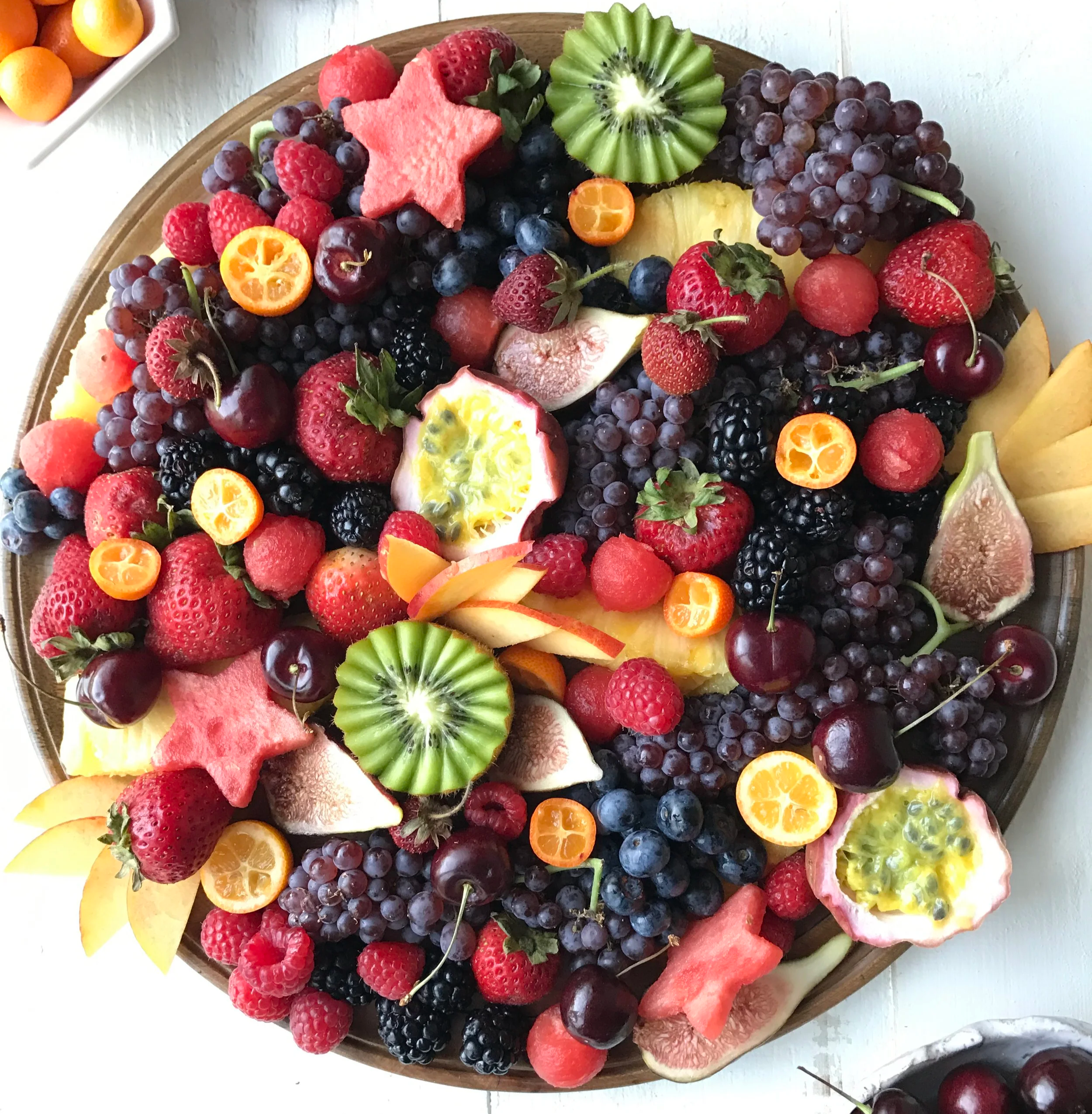 A wooden tray filled with various sliced and whole fruits including strawberries, blackberries, blueberries, red and yellow grapes, kiwi, watermelon, figs, and small orange kumquats.