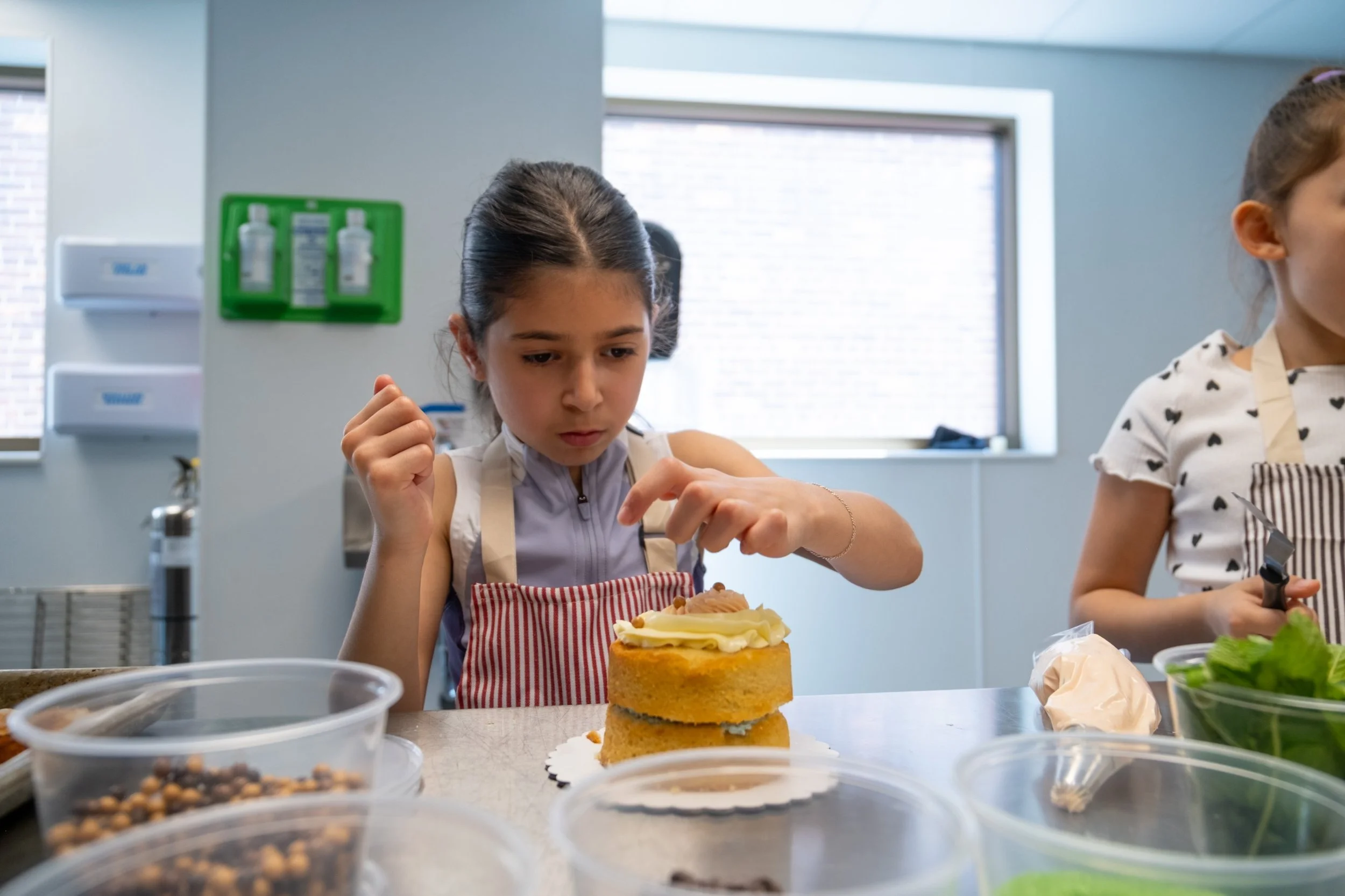 A young girl in an apron is decorating a cake with cream, surrounded by various toppings and ingredients in containers, in a kitchen or bakery setting.