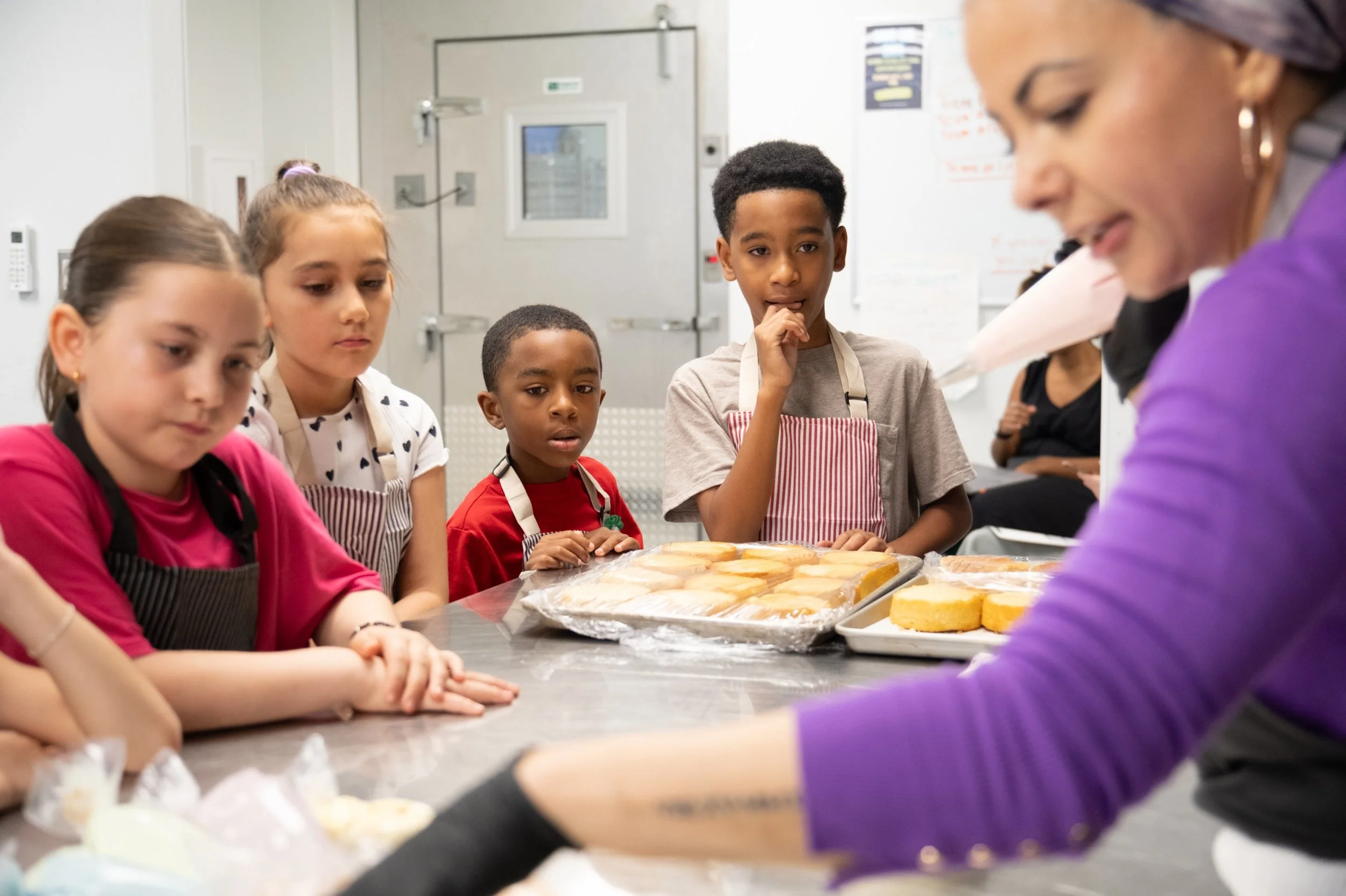 Group of children wearing aprons gathered around a table with baked goods in a kitchen or teaching environment.