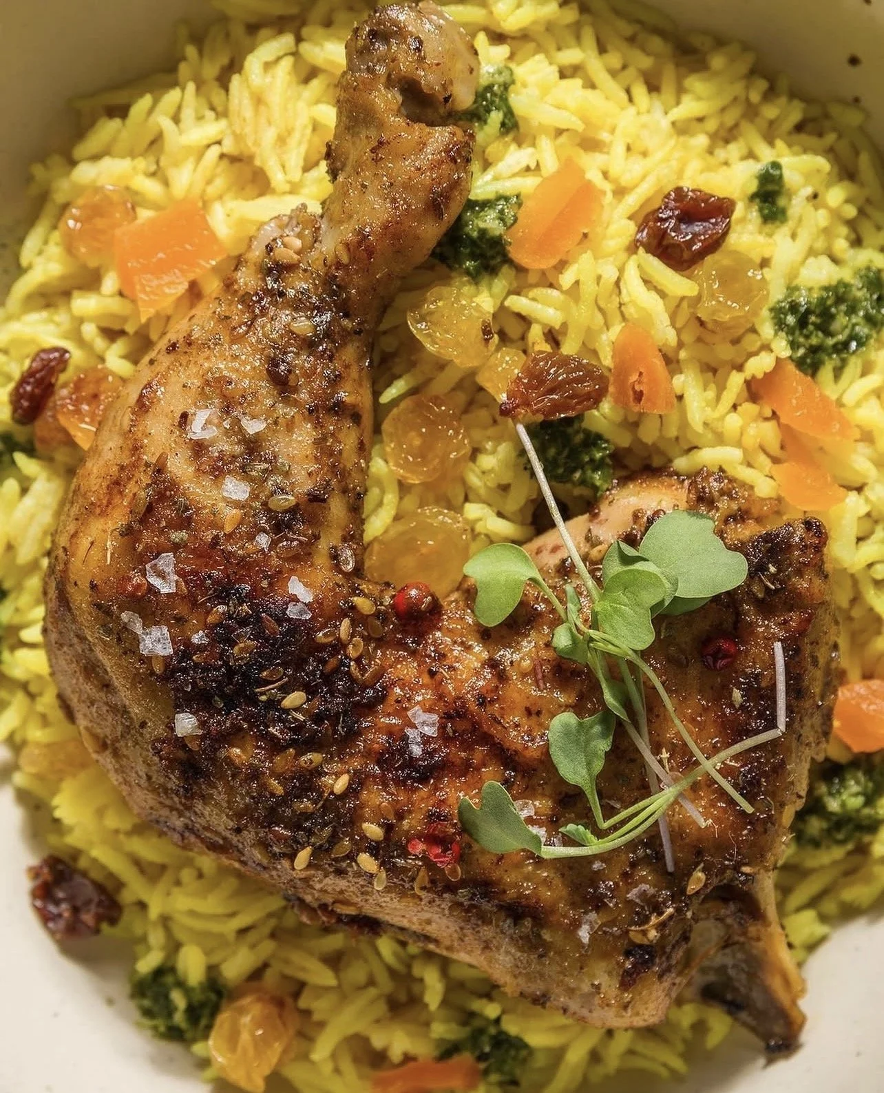 Close-up of a cooked chicken leg garnished with microgreens, served over yellow rice mixed with raisins, chopped carrots, broccoli, and cranberries.
