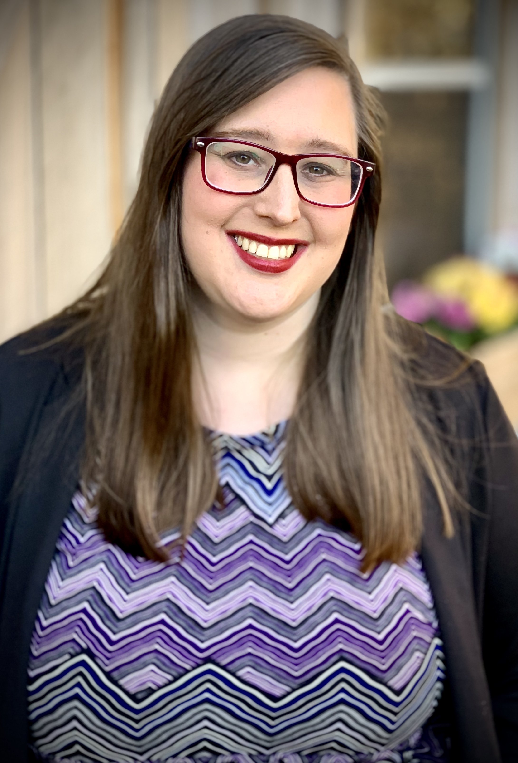 A woman with long brown hair, wearing red glasses and a patterned purple, black, and white top, smiling.