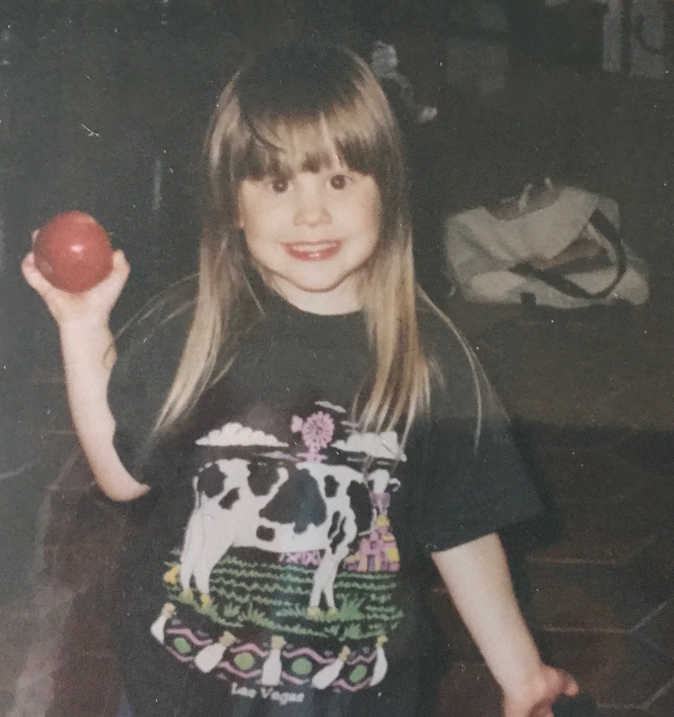 Young girl with long hair smiling, holding a red ball, wearing a black t-shirt with colorful designs, standing on a dark surface.