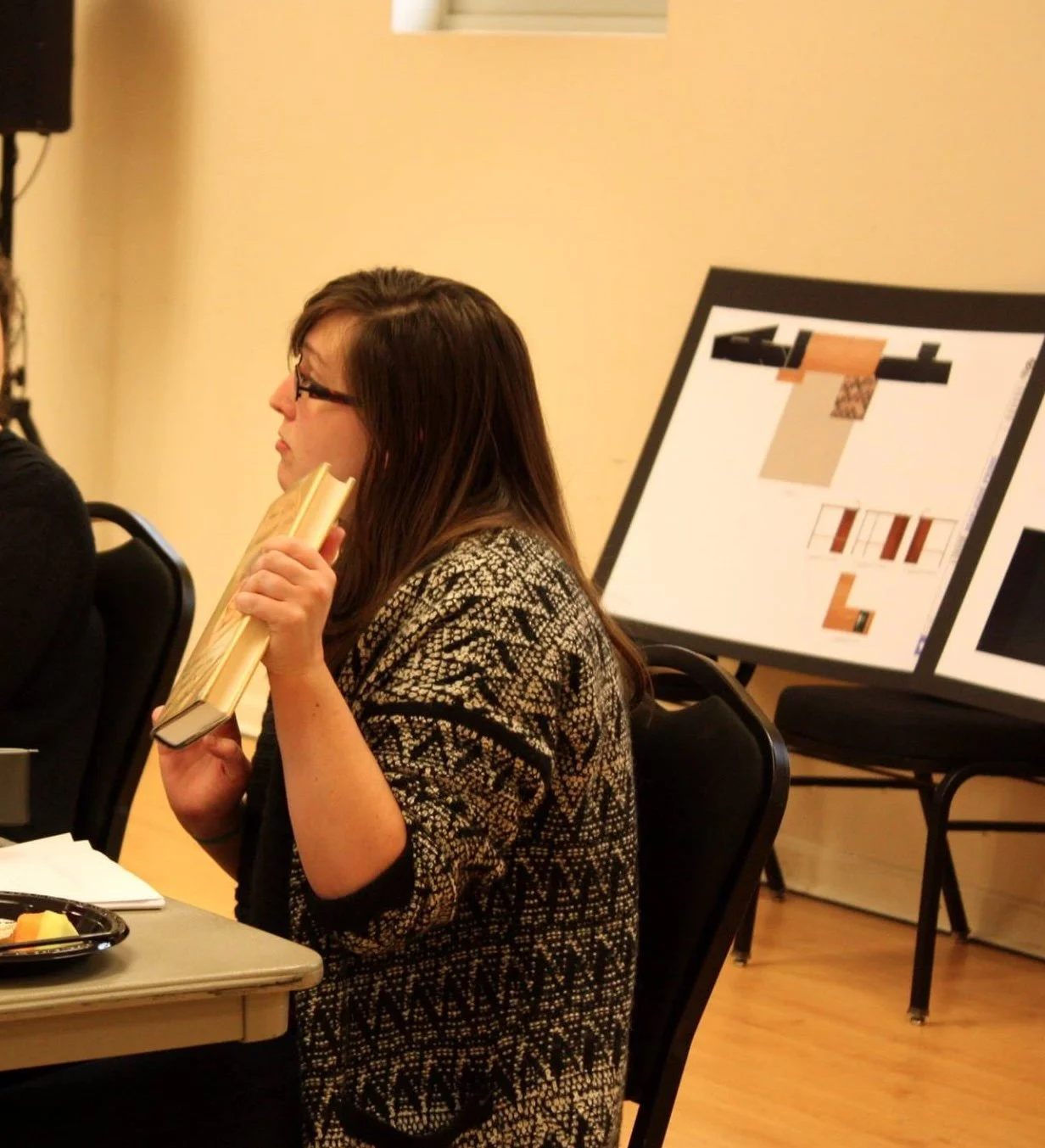A woman with glasses and long dark hair seated at a table, holding a book close to her face, with an interest in what she is reading. Behind her, a large display board shows various color swatches and patterns, indicating a design or planning session.
