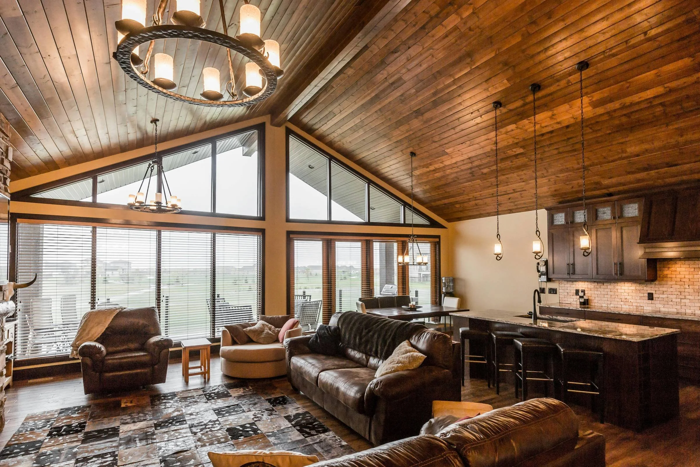 Living room with a vaulted wooden ceiling, large windows with blinds, and multiple hanging lights. Furniture includes leather sofas, a dining table, and kitchen cabinets, with a view of an outdoor patio.