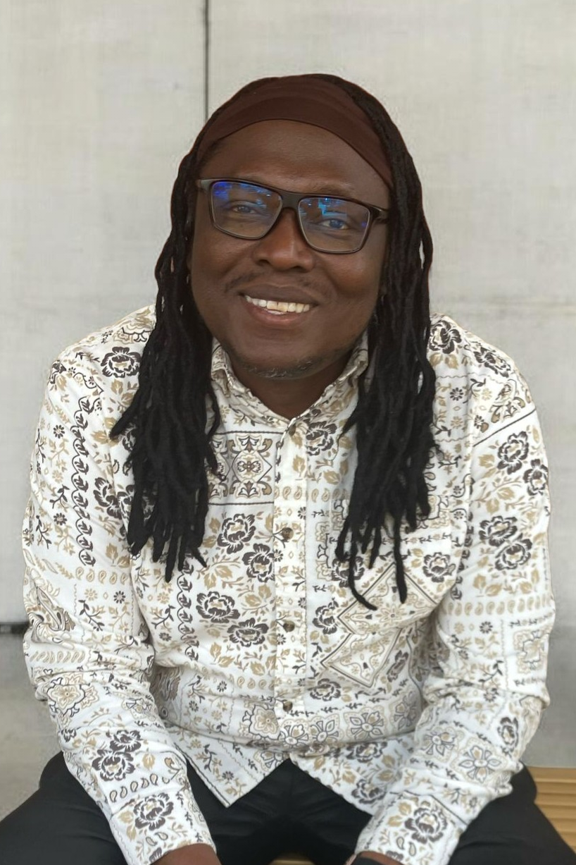 A person with long dreadlocks, glasses, and a brown headband, wearing a patterned cream, black, and gold shirt, smiling and sitting against a light-colored background.