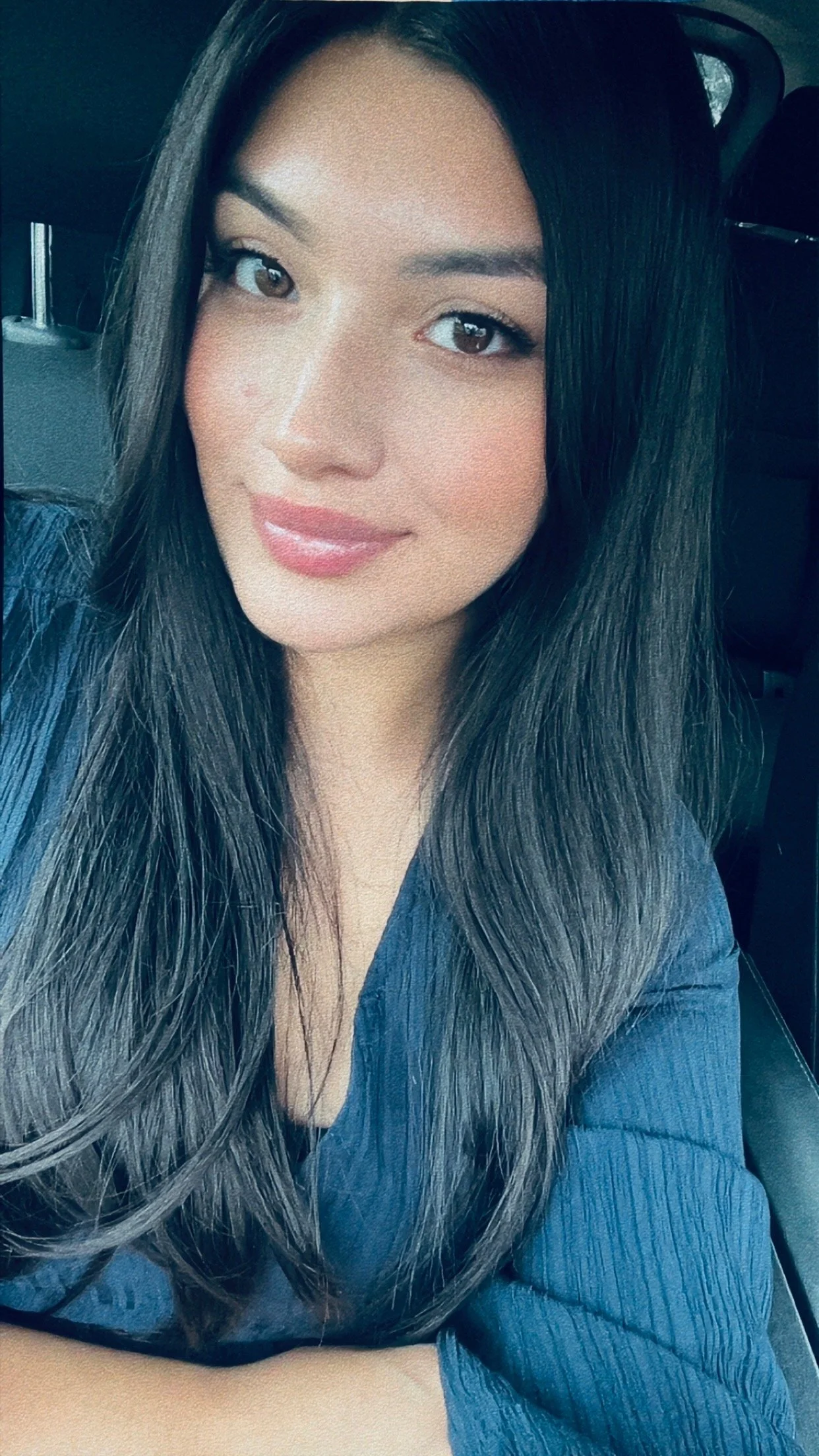 Close-up photo of a woman with long dark hair, wearing a dark blue top, smiling slightly, inside a vehicle.