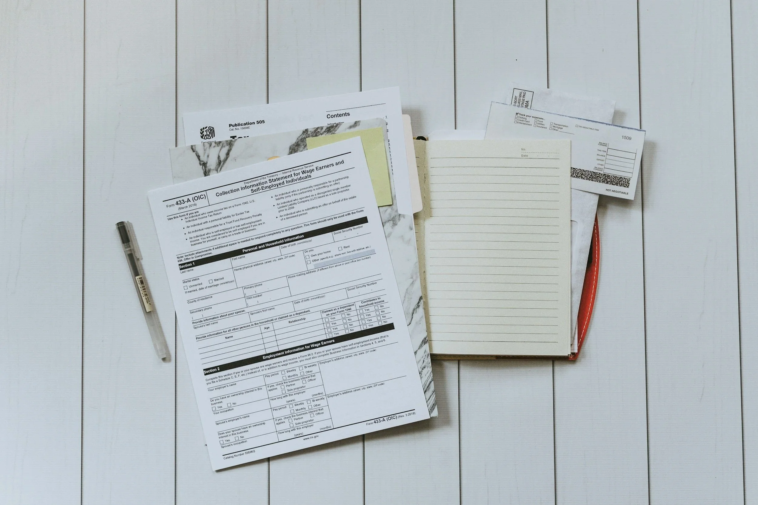 A white wooden surface with scattered papers, including a tax form, a notepad, a pen, and what appears to be a bus ticket and a checklist.