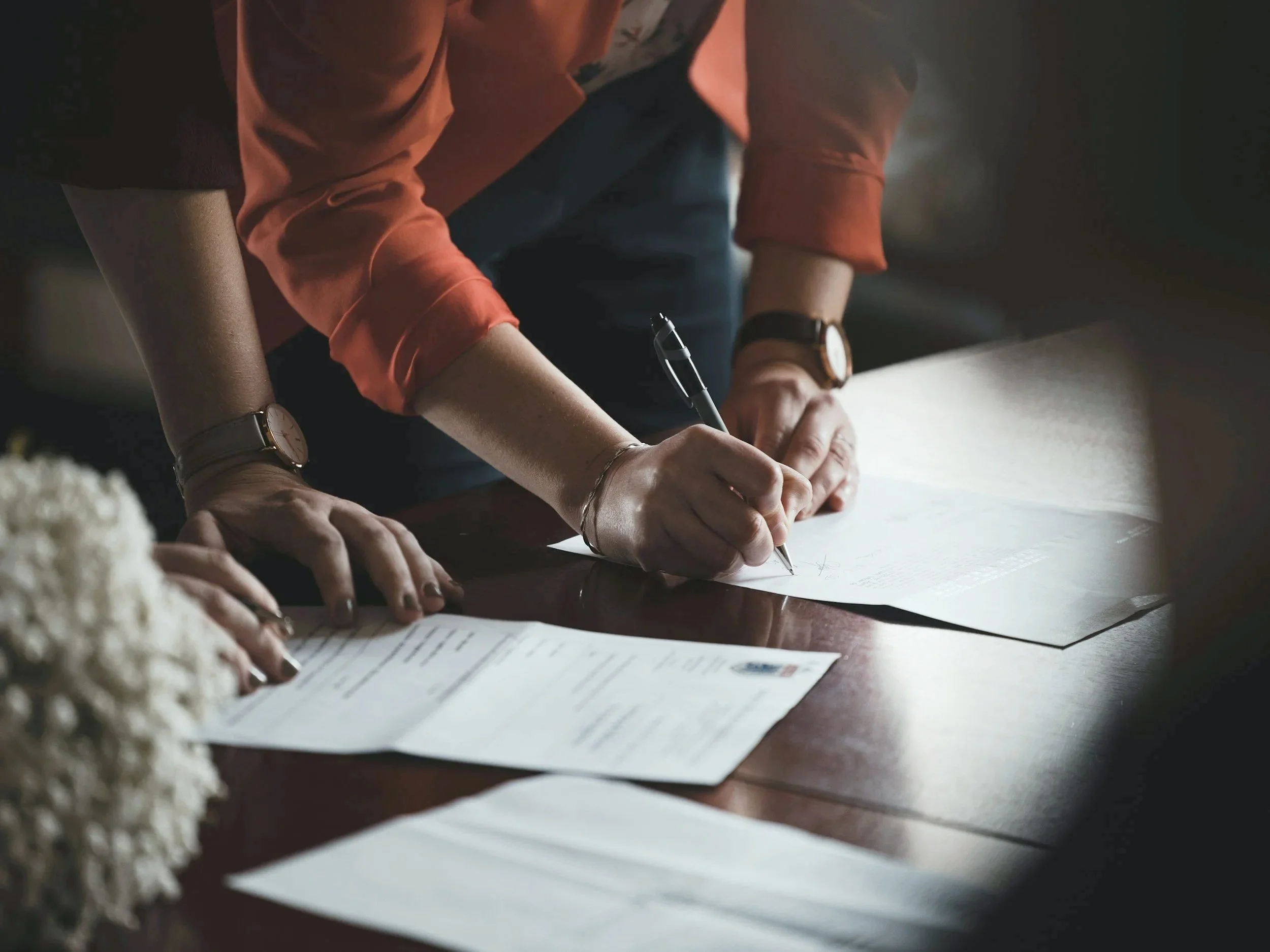 People writing on papers placed on a wooden table during a meeting or discussion, with documents and a pen visible.