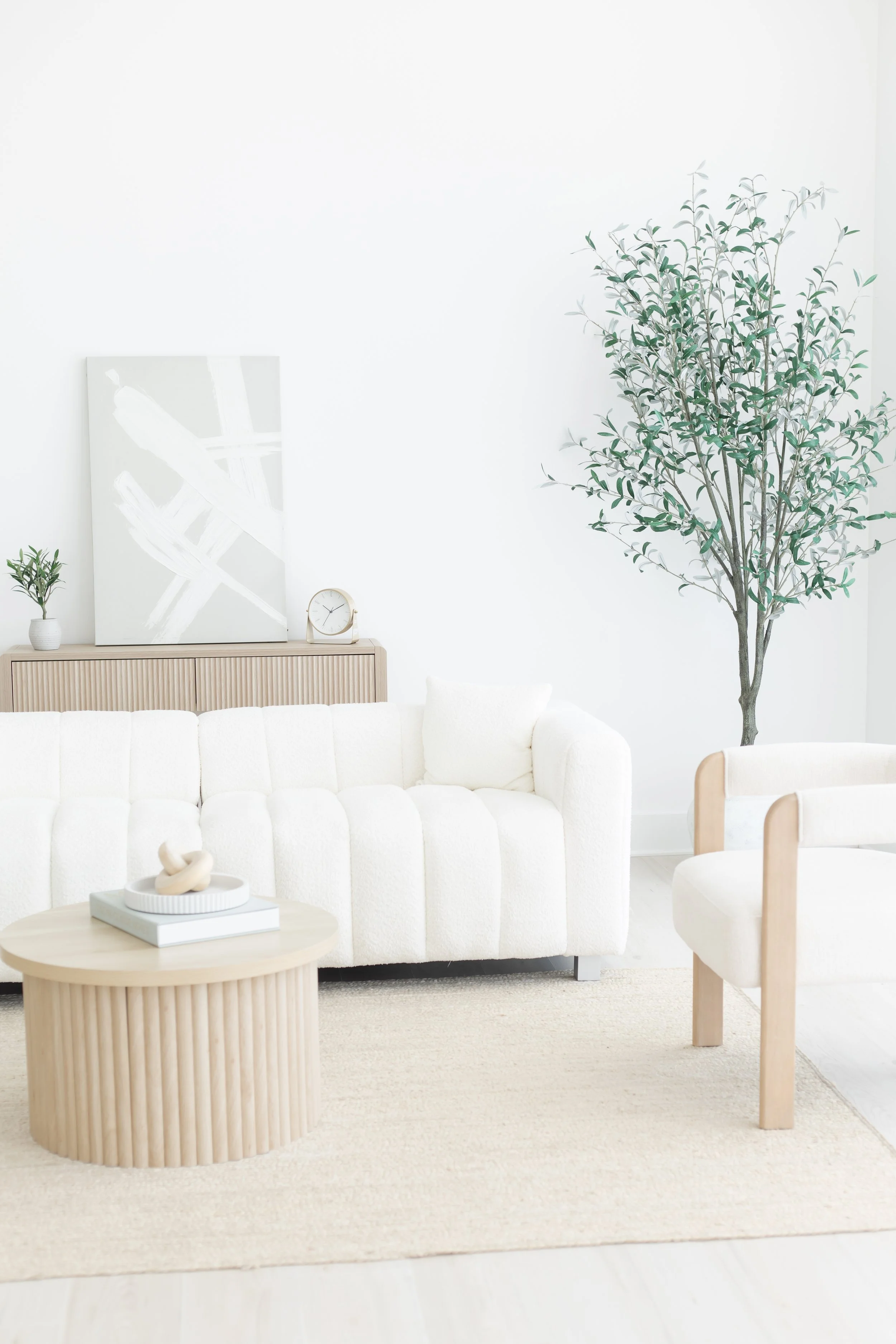 A modern minimalist living room with a white sofa, a round wooden coffee table with a white tray and abstract sculpture, a beige textured rug, a large green leafy plant on the right, and a wooden sideboard with a clock, small plant, and abstract canvas art.