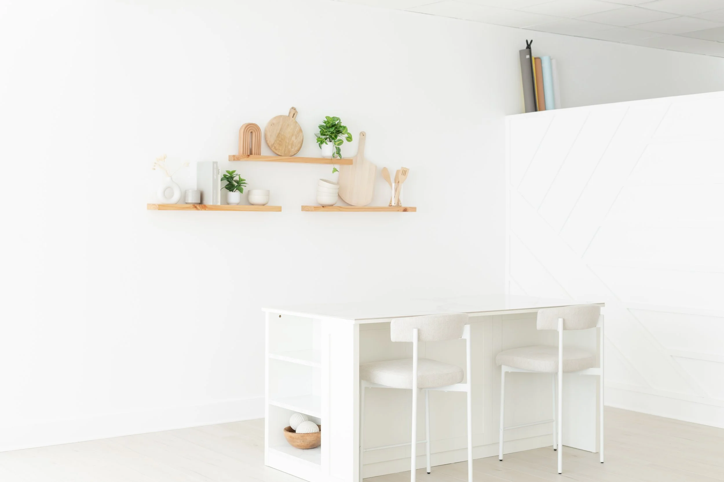 A minimalist kitchen with white walls, wooden shelves holding cutting boards, plants, bowls, and utensils, and a white dining table with chairs in the foreground.