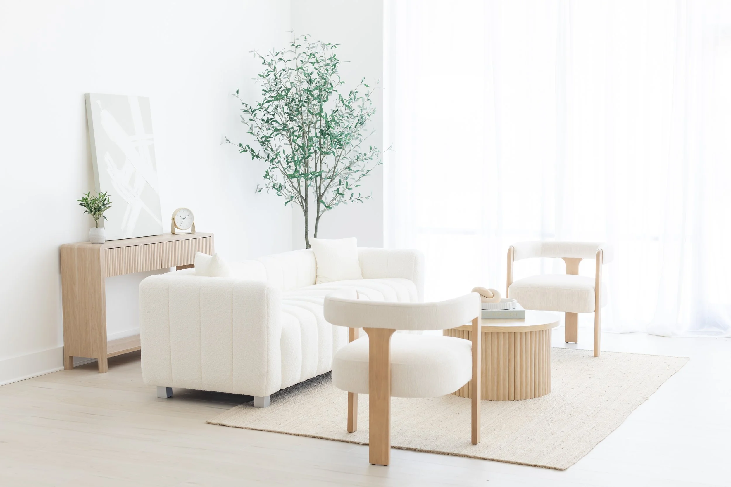Bright living room with white walls, light wood furniture, a sofa, armchair, round coffee table, large indoor tree, and sheer white curtains.