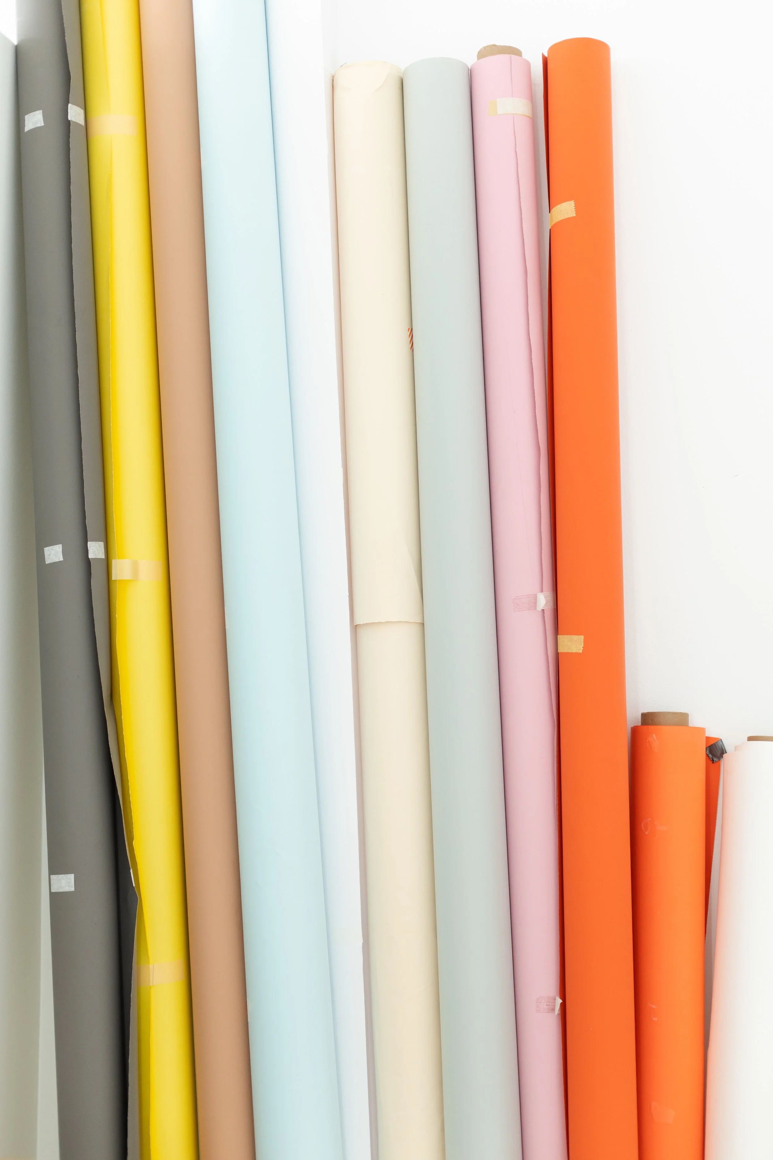 A row of colorful rolls of craft paper in shades of gray, yellow, beige, light blue, off-white, pink, orange, and brown, standing vertically.